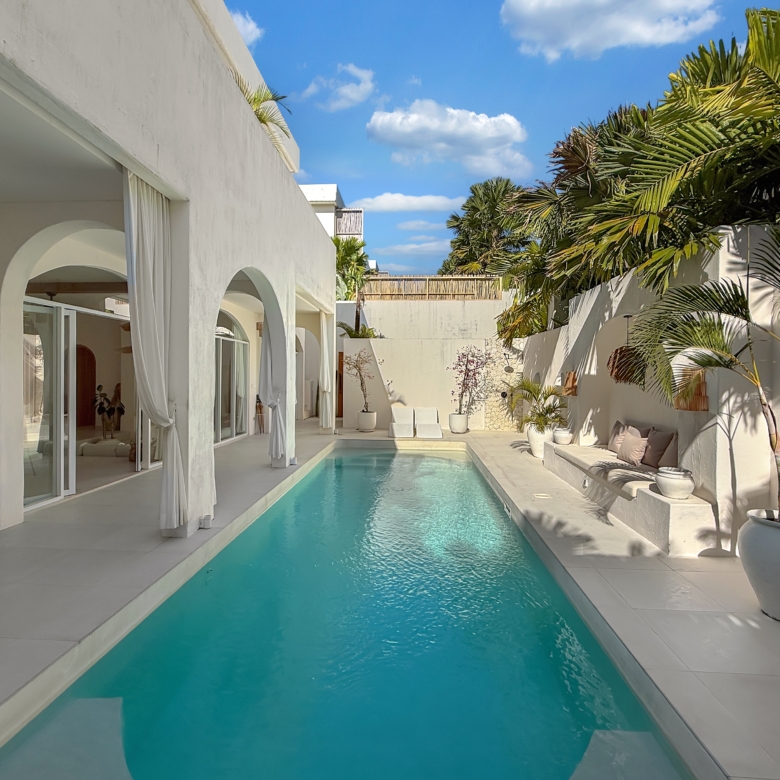 White modern villa courtyard with a rectangular turquoise pool, arches, glass doors, and draped curtains along a sunny patio.