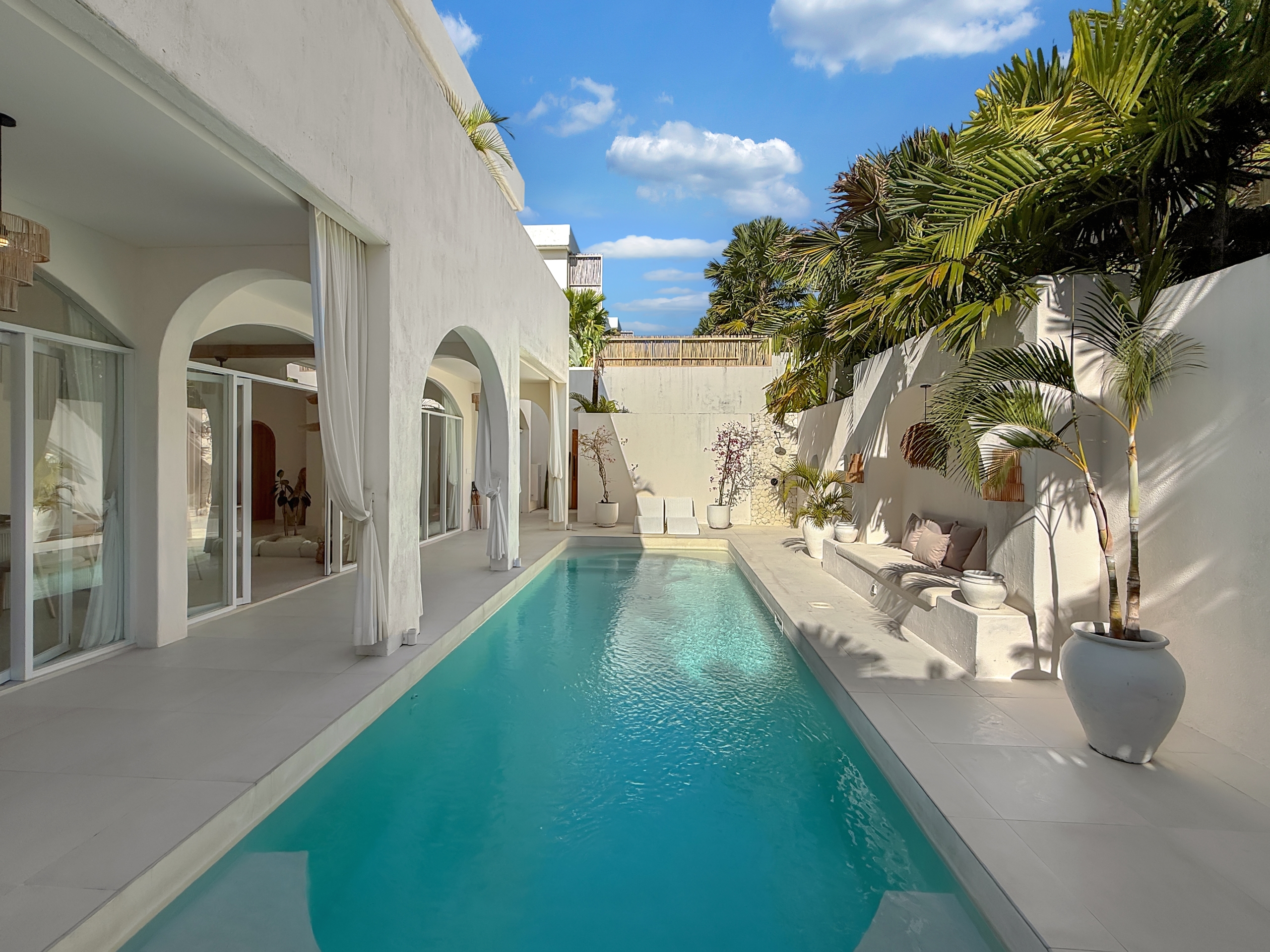 2ede1e59068c7e60776f87ba033cc1212cb9cf22 White modern villa courtyard with a rectangular turquoise pool, arches, glass doors, and draped curtains along a sunny patio.