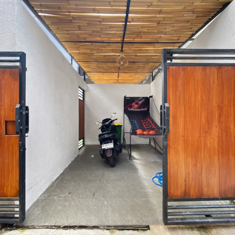 Open wrought-iron gate leading to a narrow covered passage with wooden doors, a scooter parked inside, and a wicker lantern hanging from a wooden slatted ceiling.