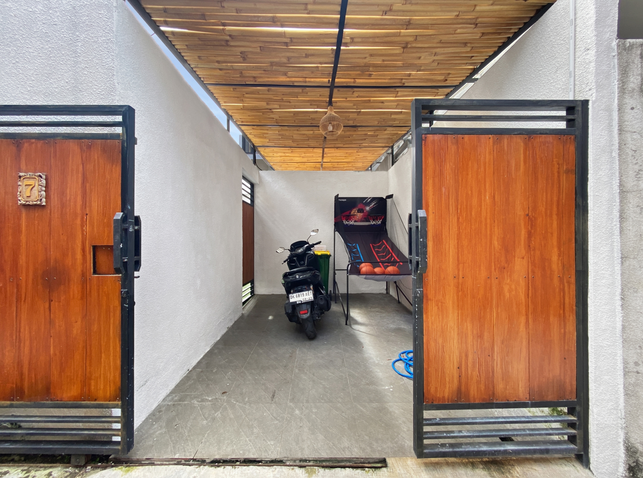 2f97f68e64fce7d72b0b67e9ed00542b67afb0cd Open wrought-iron gate leading to a narrow covered passage with wooden doors, a scooter parked inside, and a wicker lantern hanging from a wooden slatted ceiling.