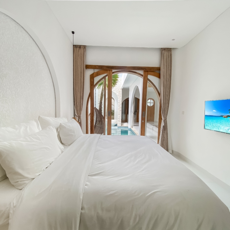 Bright hotel bedroom with white bedding, curved wall, and a wooden arched doorway leading to an outdoor pool area; a TV shows a tropical beach.