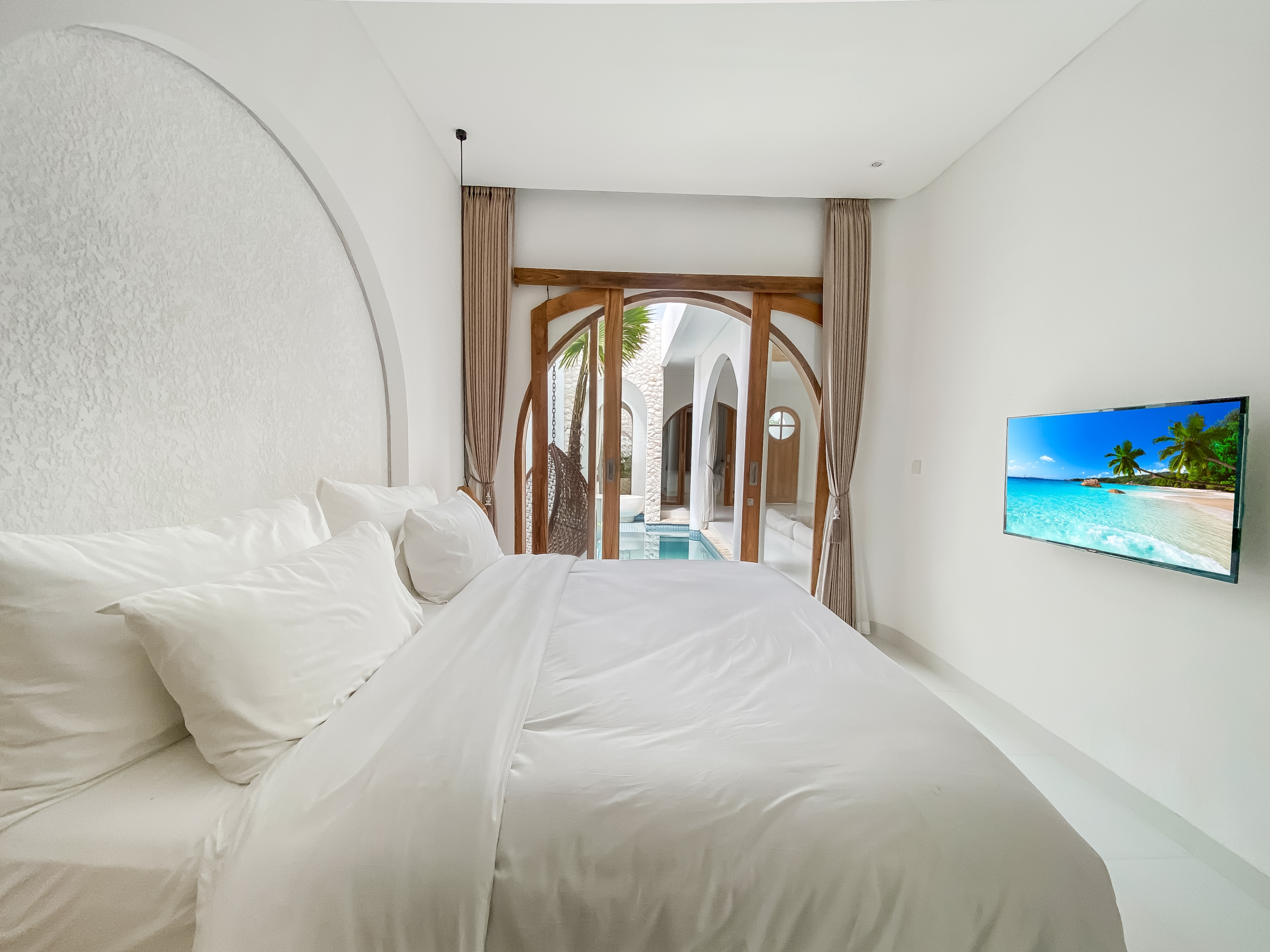 30a81fdf3f8cbe4680b158f6ca21cd956784a408 Bright hotel bedroom with white bedding, curved wall, and a wooden arched doorway leading to an outdoor pool area; a TV shows a tropical beach.