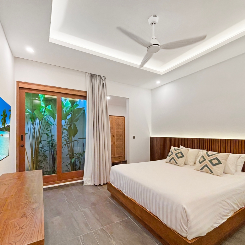 Hotel bedroom with white linen bed and wooden headboard, wall TV on the left, and sliding glass doors to greenery outside.