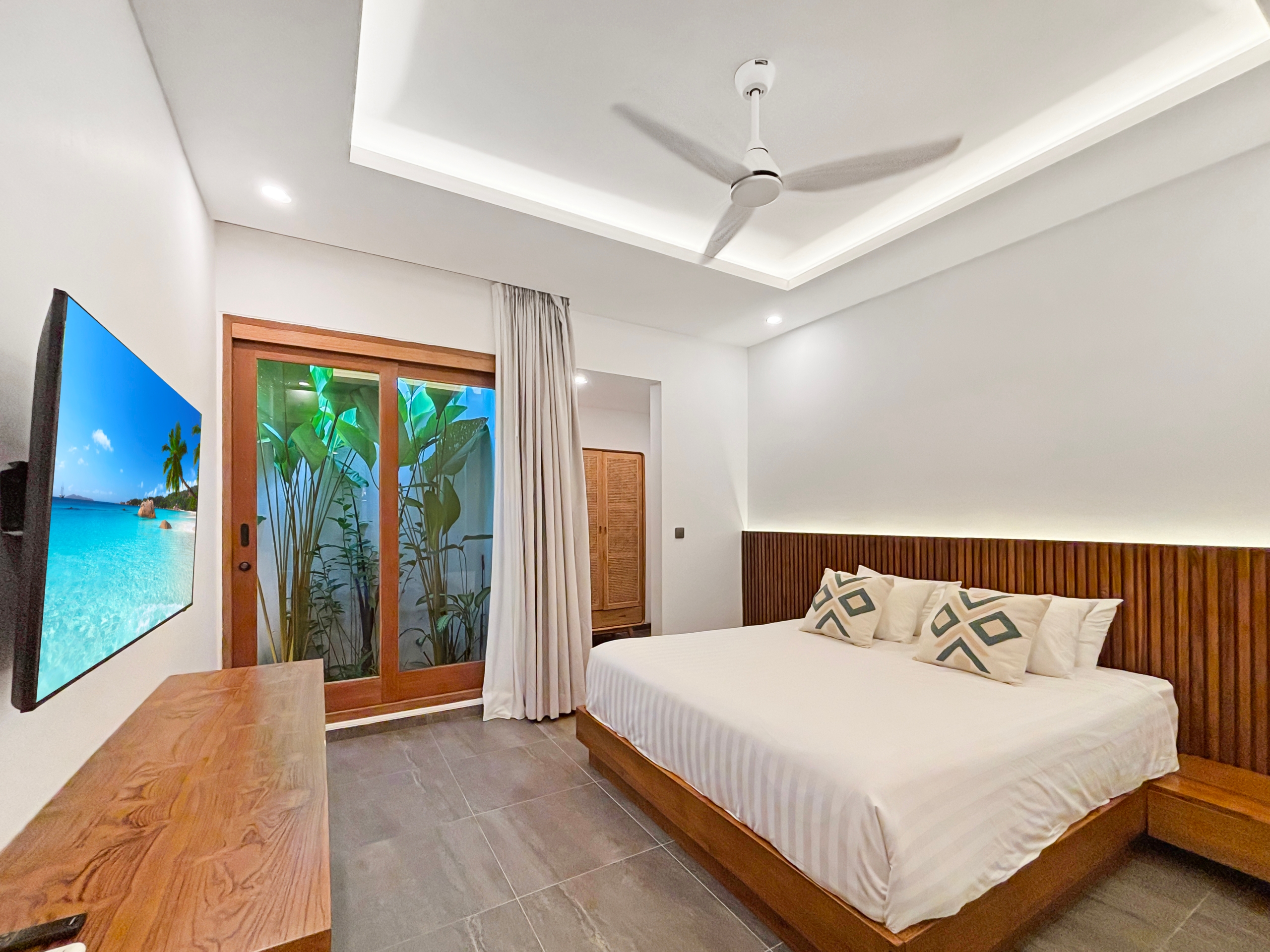 3651758e9a5fac5eb703b831e39921927669dfcc Hotel bedroom with white linen bed and wooden headboard, wall TV on the left, and sliding glass doors to greenery outside.