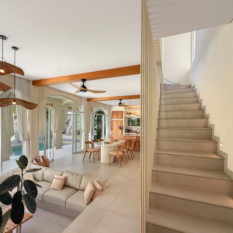 Bright open-plan living area with a beige sectional, white floors, and large glass walls overlooking a pool; staircase on the right and wicker pendant lights above.