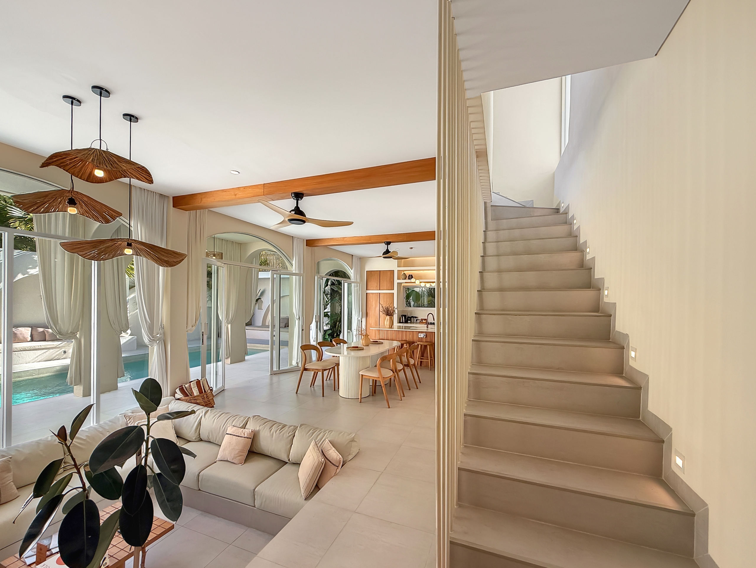 36c37c44c26146f73abe7bdb857ca6728569fb80 Bright open-plan living area with a beige sectional, white floors, and large glass walls overlooking a pool; staircase on the right and wicker pendant lights above.