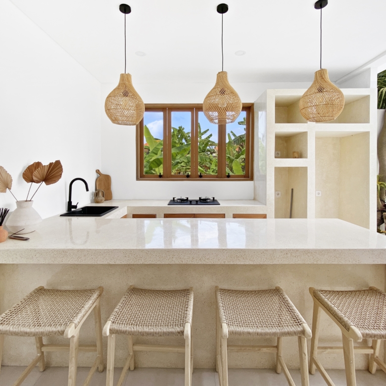 Kitchen counter with four woven-straw stools, three hanging wicker pendant lights, and a window view of tropical plants outside.