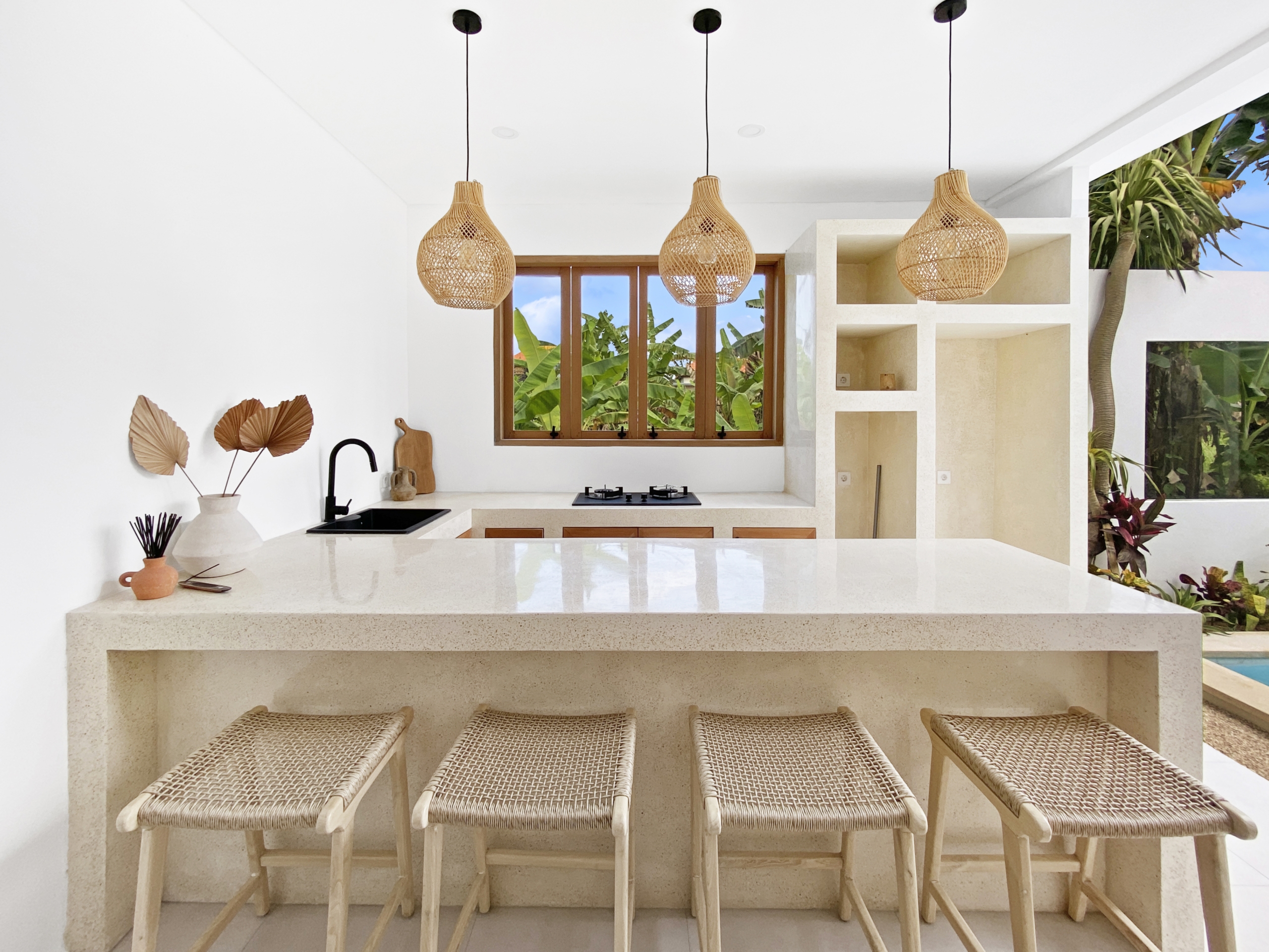 38fdc1db0e1ab4a1f39990a9d79ea0e31528d04f_2314-68d5cfb233b61 Kitchen counter with four woven-straw stools, three hanging wicker pendant lights, and a window view of tropical plants outside.