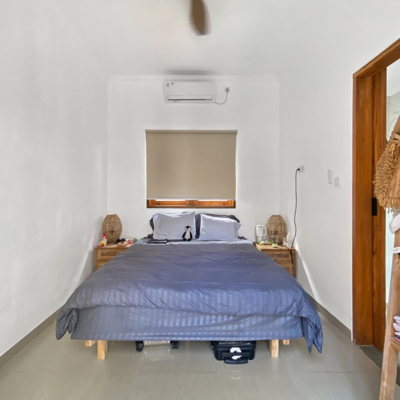 Bedroom with a centered bed in blue-gray bedding, air conditioner above the window, and wooden shelving on the right.