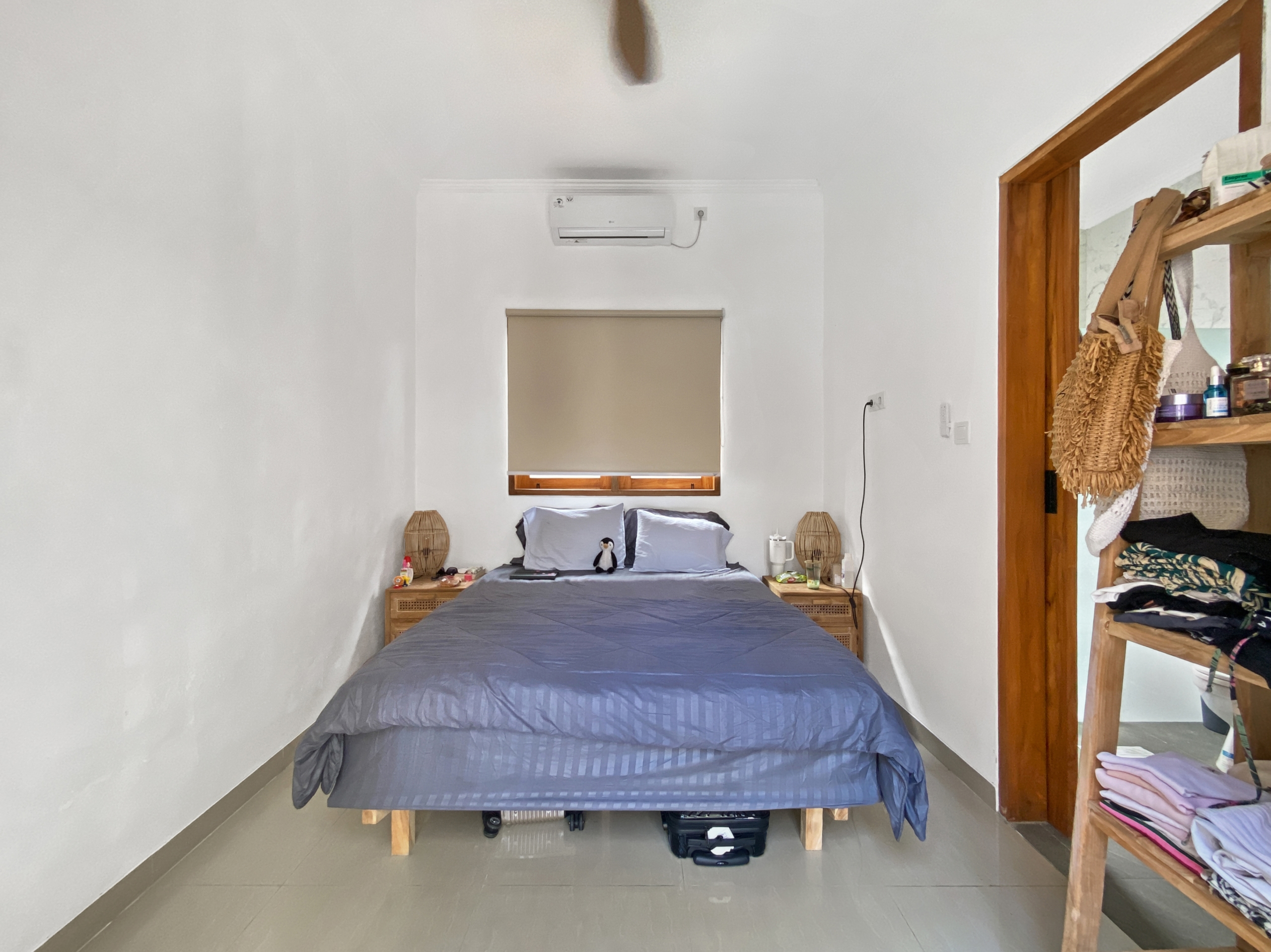 394d6c522818b0368e1be560f441fb0601493042 Bedroom with a centered bed in blue-gray bedding, air conditioner above the window, and wooden shelving on the right.