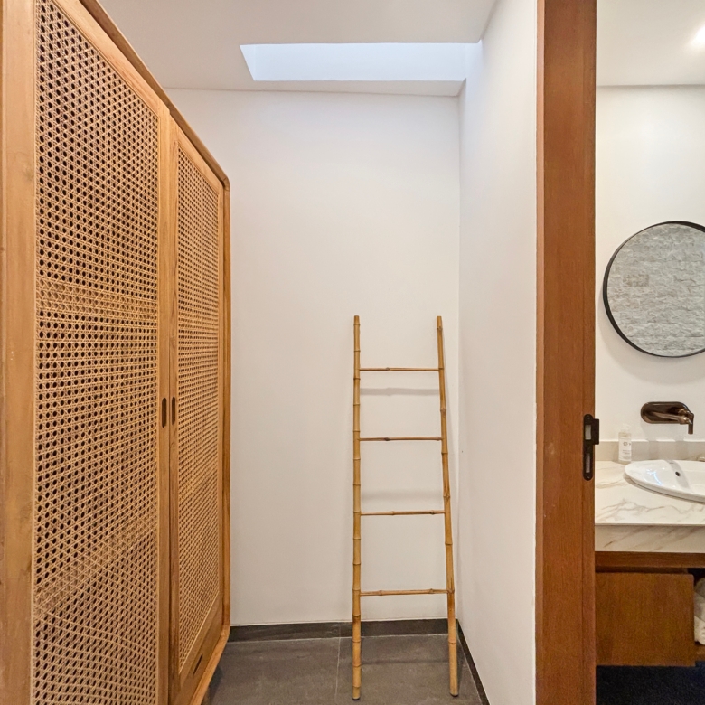 Wooden cabinet with woven panels on the left, a tall bamboo ladder in the center, and a round wall mirror on the right.