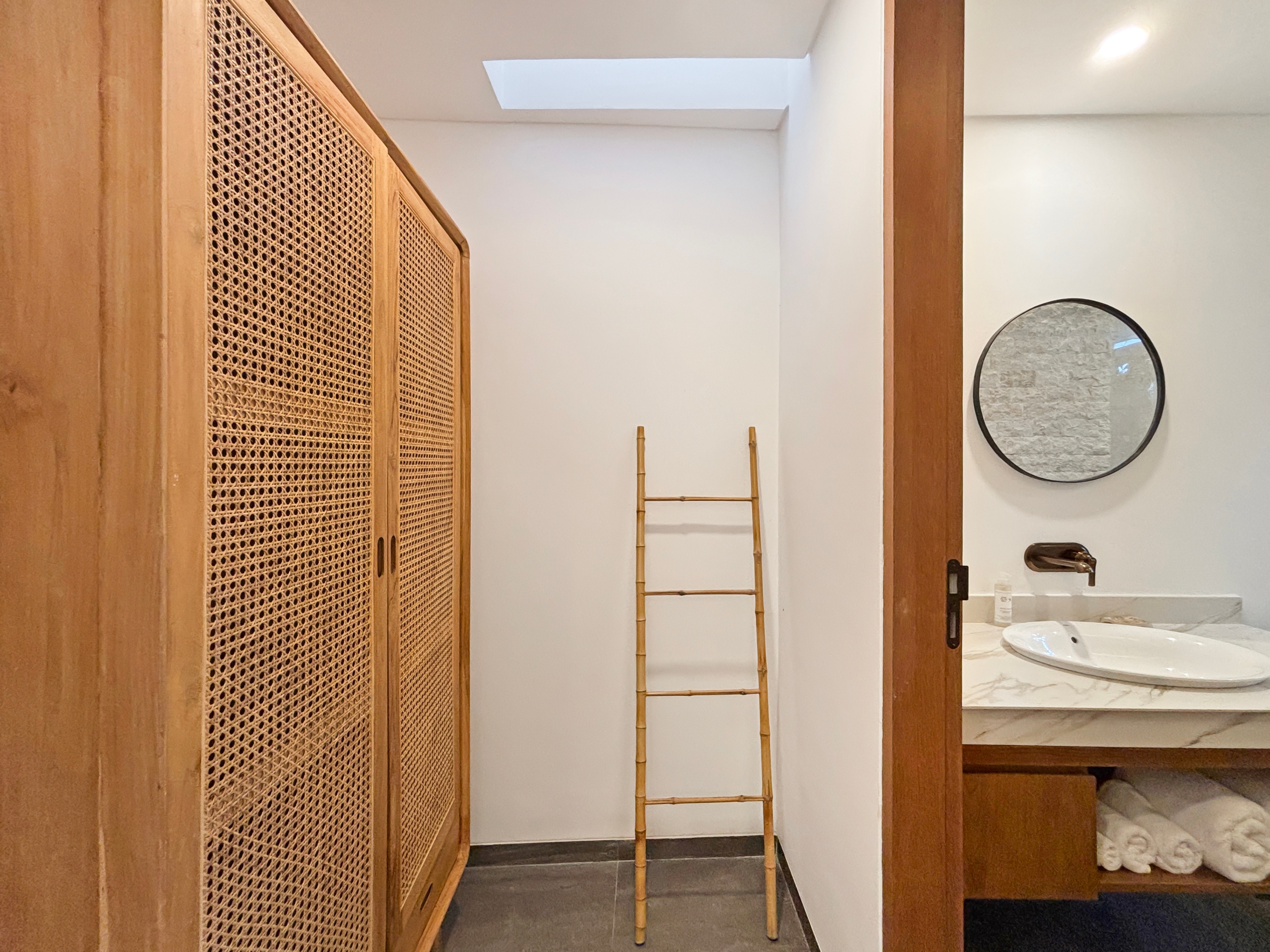 3b4c2eea227d7a4128f273a9c554c59a762aa7a7 Wooden cabinet with woven panels on the left, a tall bamboo ladder in the center, and a round wall mirror on the right.