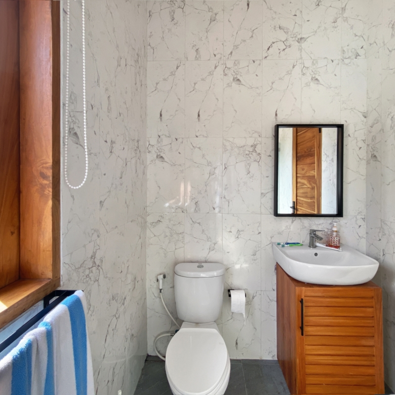 Bathroom with marble walls, wooden window frame, and a sink with a wooden vanity opposite a toilet; a mirror on the wall and a blue-and-white towel draped over a radiator.