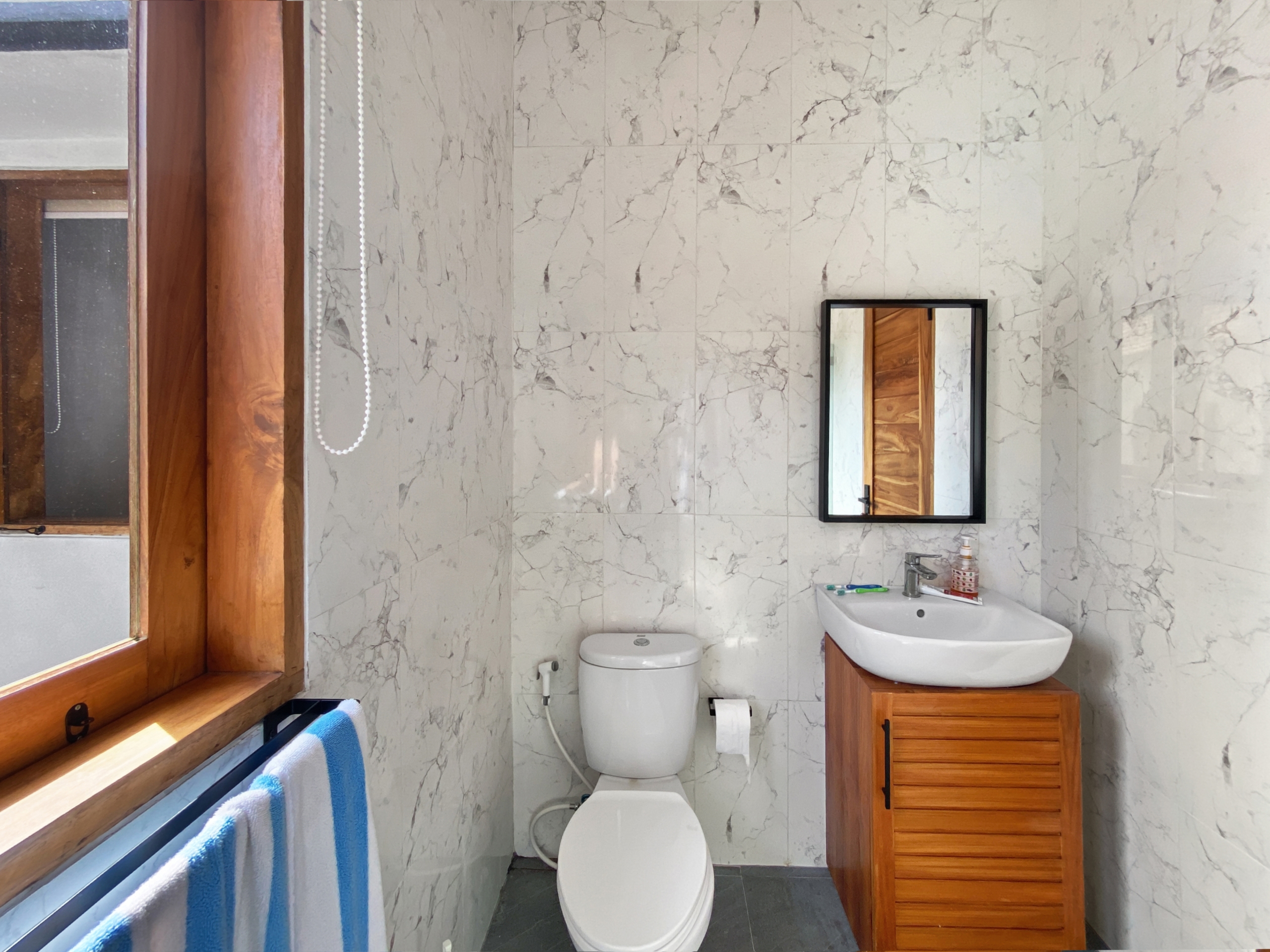 3c7781532f98924c2530d3c618604637b9971f06 Bathroom with marble walls, wooden window frame, and a sink with a wooden vanity opposite a toilet; a mirror on the wall and a blue-and-white towel draped over a radiator.