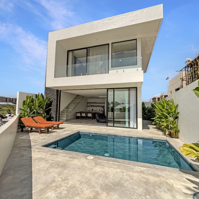 Modern white multi‑level house with large glass walls and a rectangular swimming pool in the foreground under a clear blue sky.