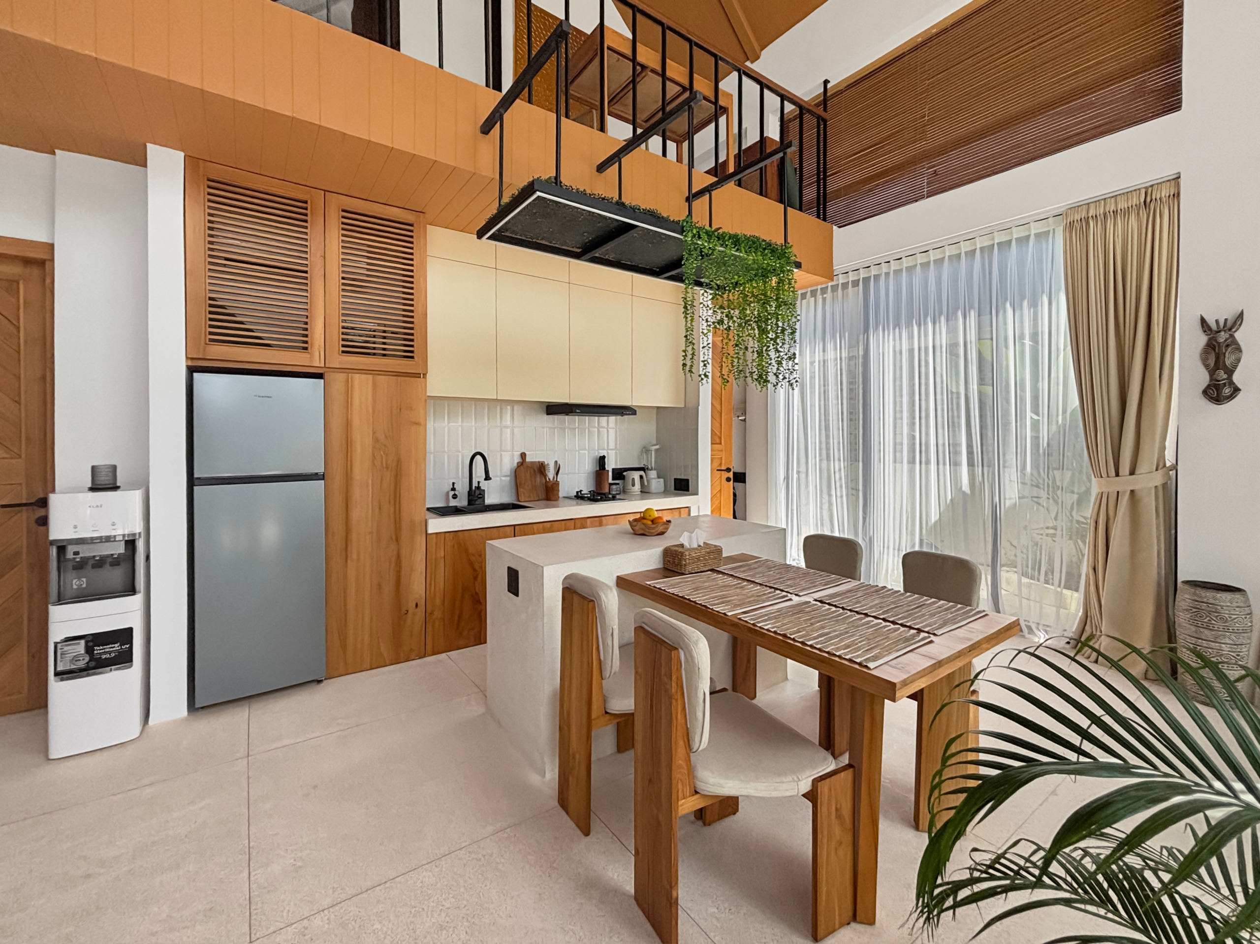 3df9569a57f97fbc6f905aef579a30e8f7397490 Modern kitchen with wooden cabinets, stainless fridge, and a white island; a hanging planter shelf with greenery over the island and a dining table nearby.