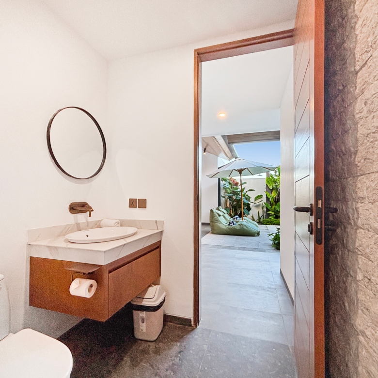 Bathroom vanity with round mirror, sink, and a toilet; a wooden door is open to an outdoor patio with plants and umbrellas beyond the doorway.