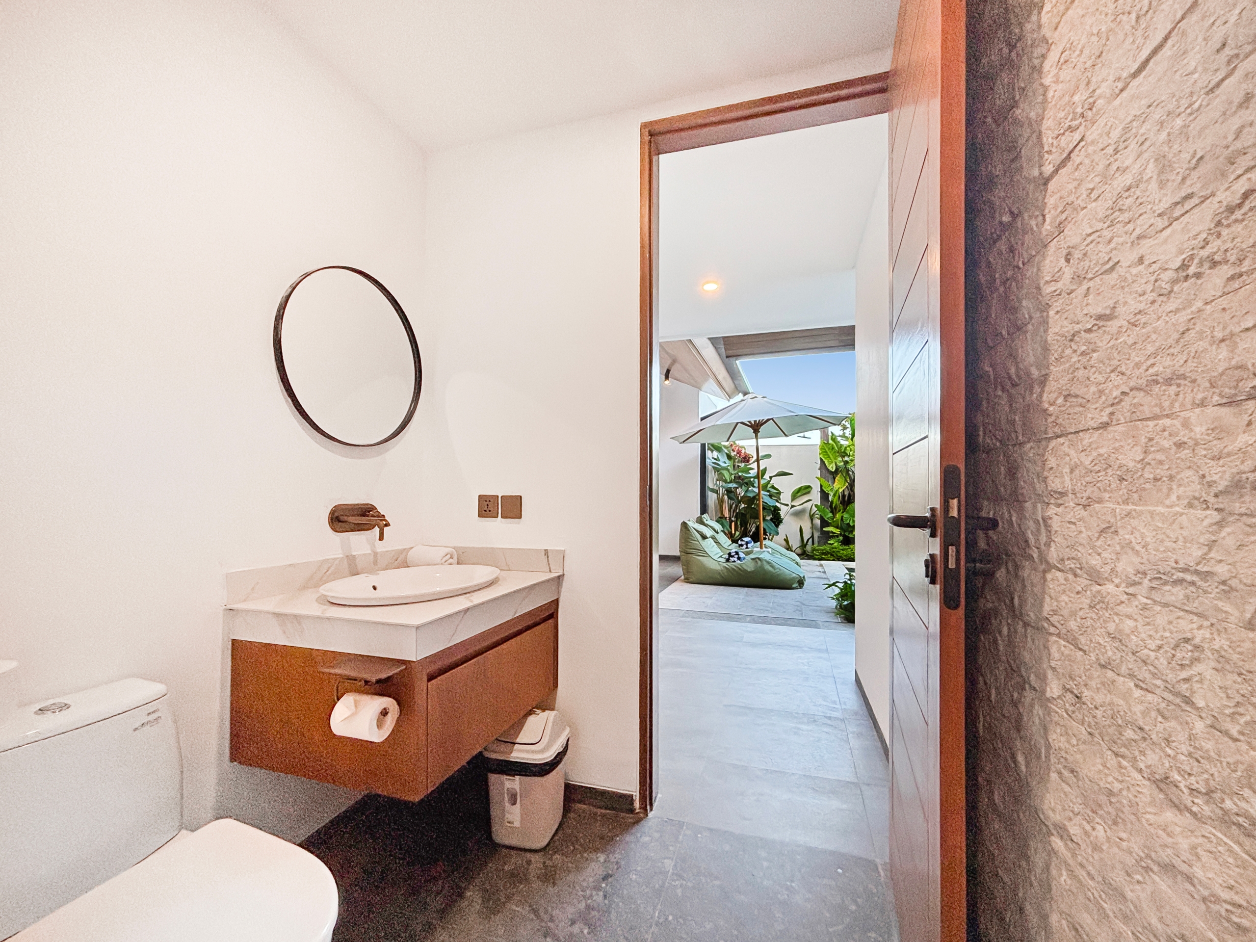 3e7ec16dc18d04c16806d895c4e7c73e0cd19bed Bathroom vanity with round mirror, sink, and a toilet; a wooden door is open to an outdoor patio with plants and umbrellas beyond the doorway.