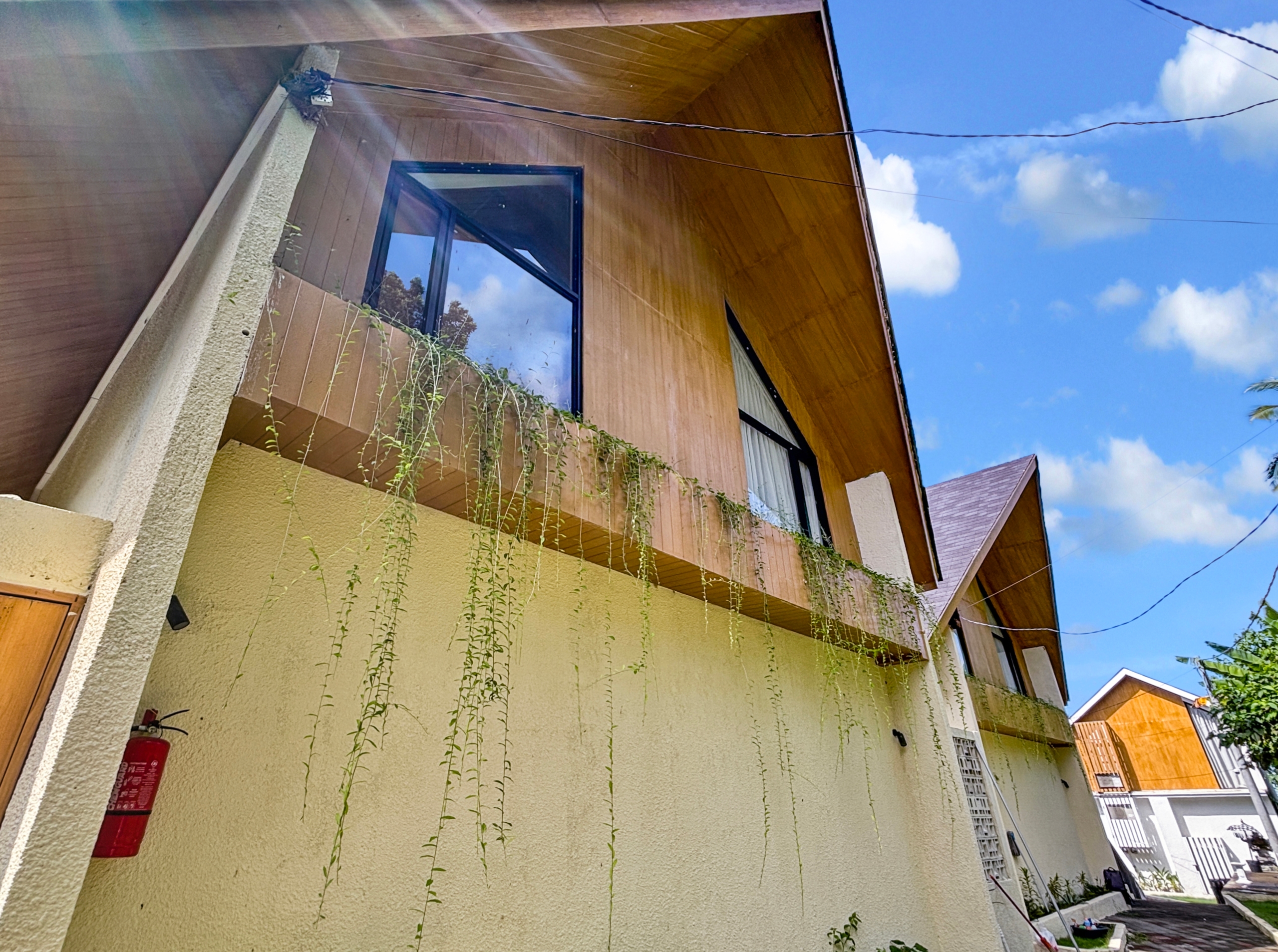 3f437f6f91474b082140e3bd0527a803ff571786 Two-story house with brown wooden siding, a large glass window, and long vines hanging down a beige stucco wall against a bright blue sky.