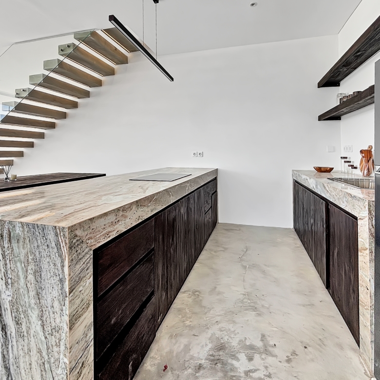 Modern kitchen with marble island, dark wood cabinets, and a glass-railed floating staircase along the left wall.