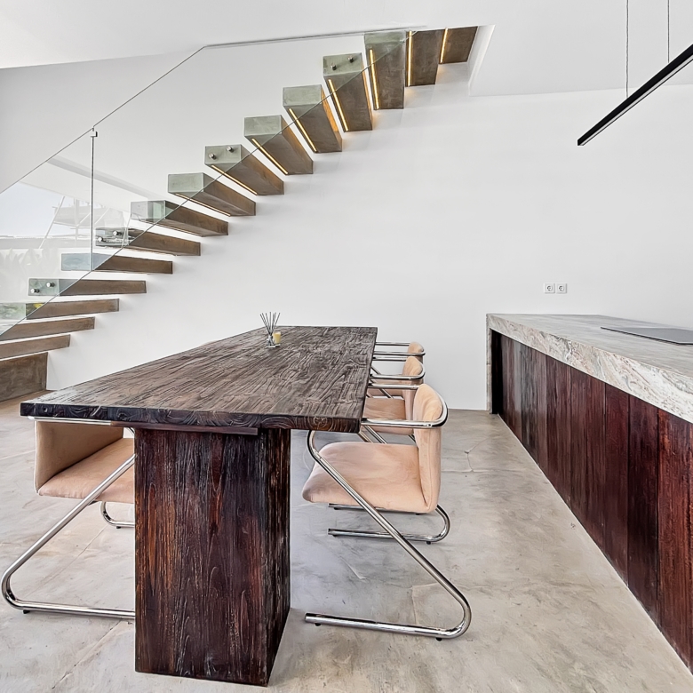 Modern interior with a diagonal floating wooden staircase and glass railing beside a large dark wood dining table and tan chairs; bright and minimal.