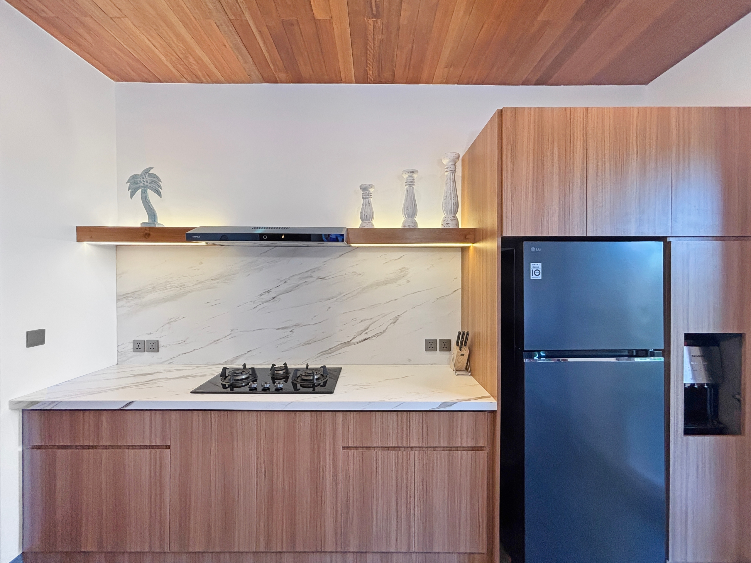 4a2f9e92f65161c51b9e35d72b352b9cc05d3bcf Modern kitchen with marble countertop, gas stove, and wooden cabinets; a shelf displays a palm statue and three white decorative pillars.
