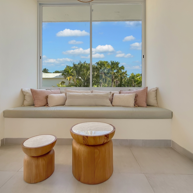 Sunlit seating nook with a built-in bench and beige cushions facing a large window framed by white walls and palm trees outside.
