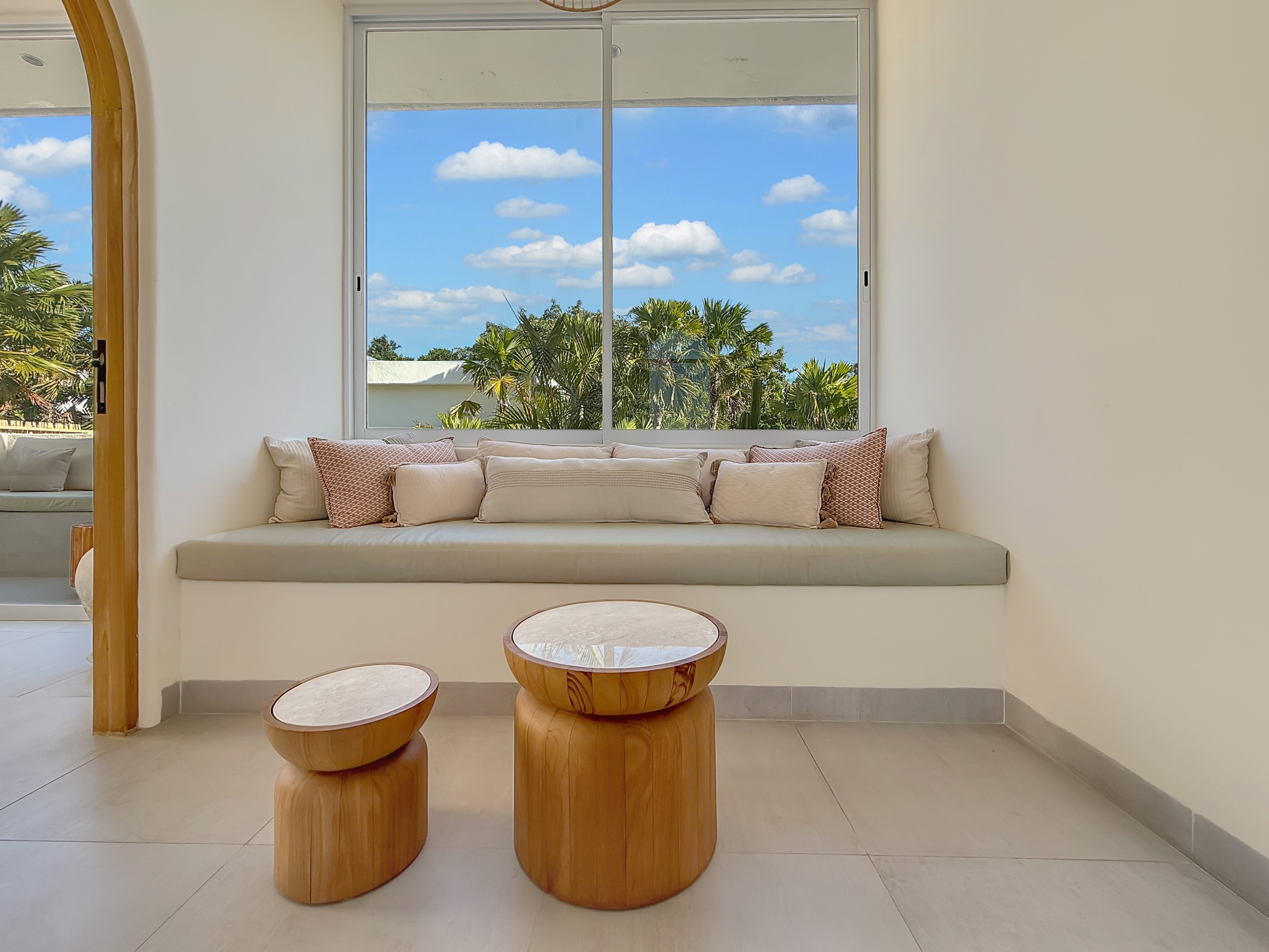 4d326c8997559bddd79fec6e38c027d61b3aa538 Sunlit seating nook with a built-in bench and beige cushions facing a large window framed by white walls and palm trees outside.