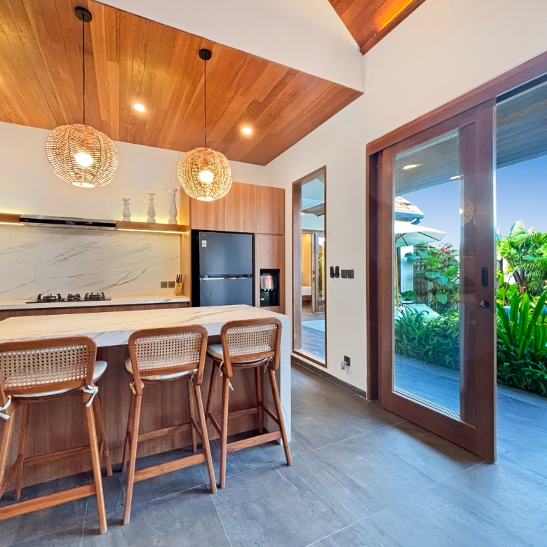 Modern kitchen with marble island, wooden bar stools, and two wicker pendant lights; glass doors open to a tropical garden outside.