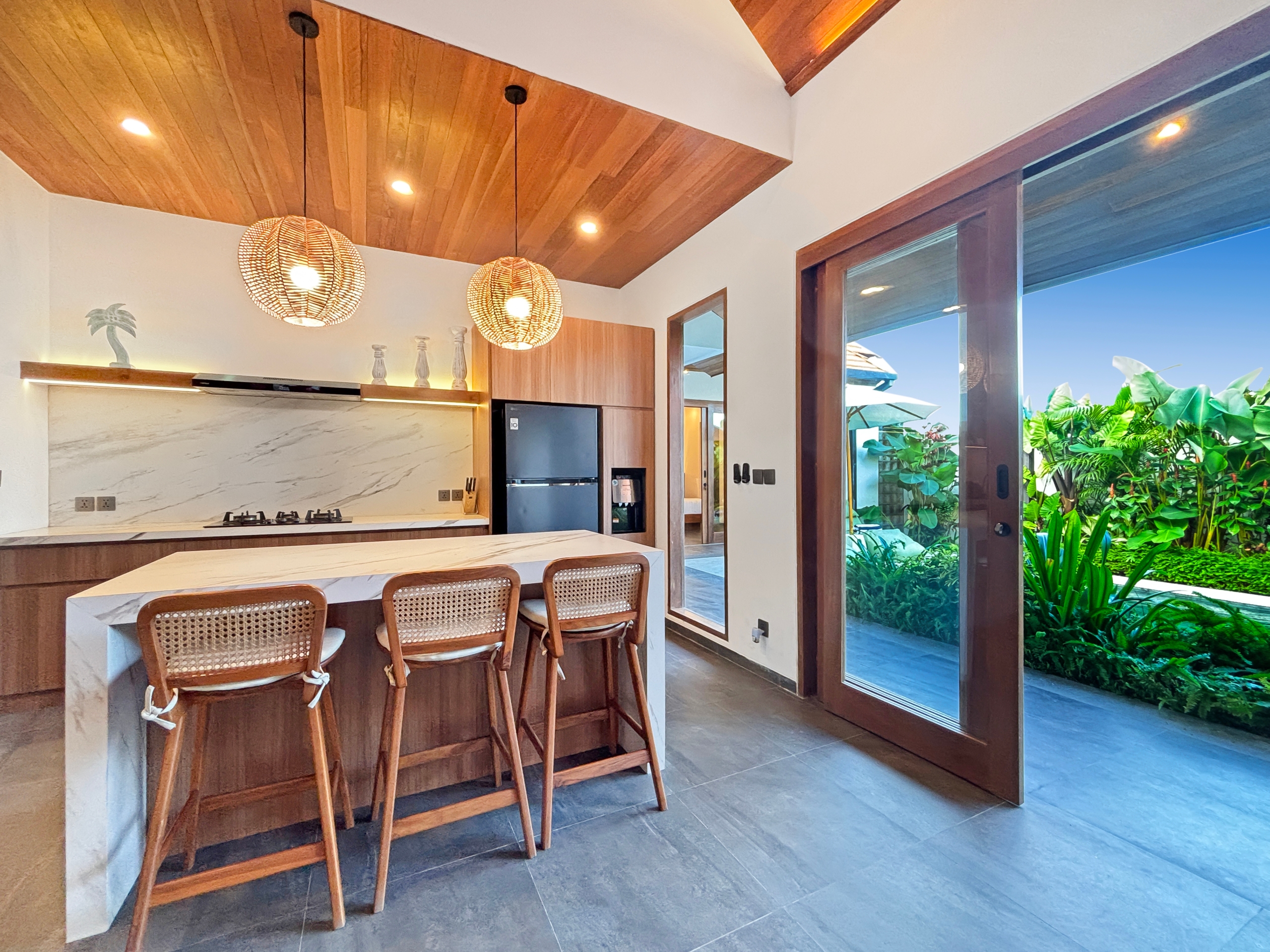 4e6d8b318792e58f755e8575dca804ed64944d2f Modern kitchen with marble island, wooden bar stools, and two wicker pendant lights; glass doors open to a tropical garden outside.