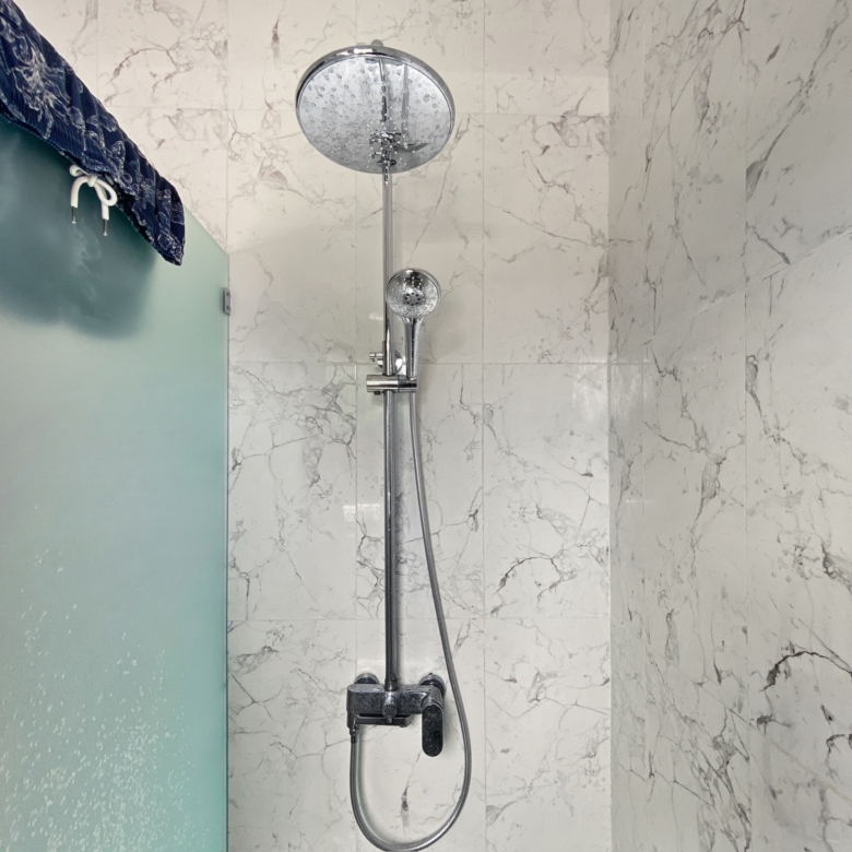Marble-tiled shower with a chrome overhead rain shower and a handheld shower on a vertical rail one a frosted glass door to the left