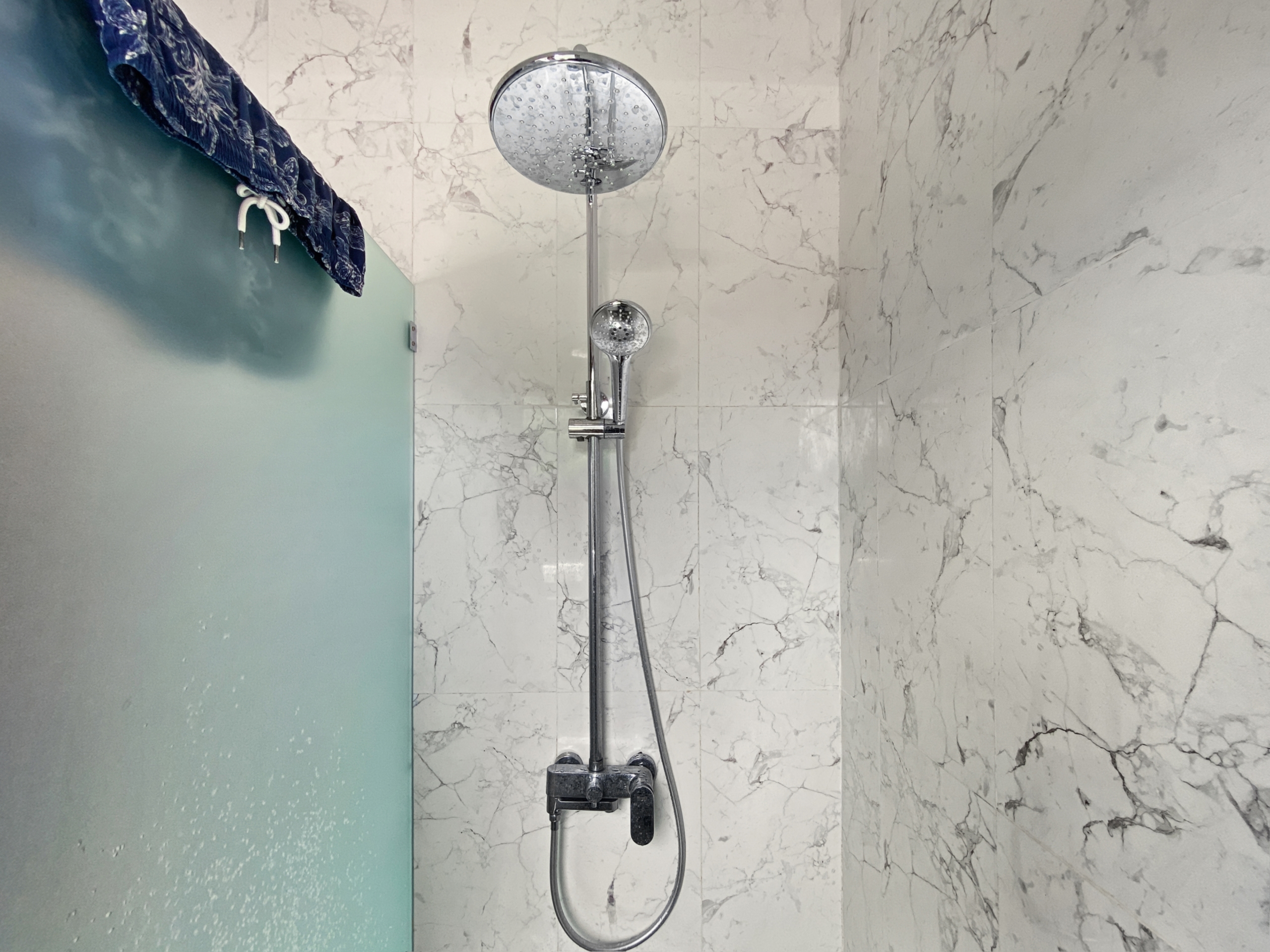 4fa075721cb612c83f1a79ee57047f81aee6a718 Marble-tiled shower with a chrome overhead rain shower and a handheld shower on a vertical rail one a frosted glass door to the left