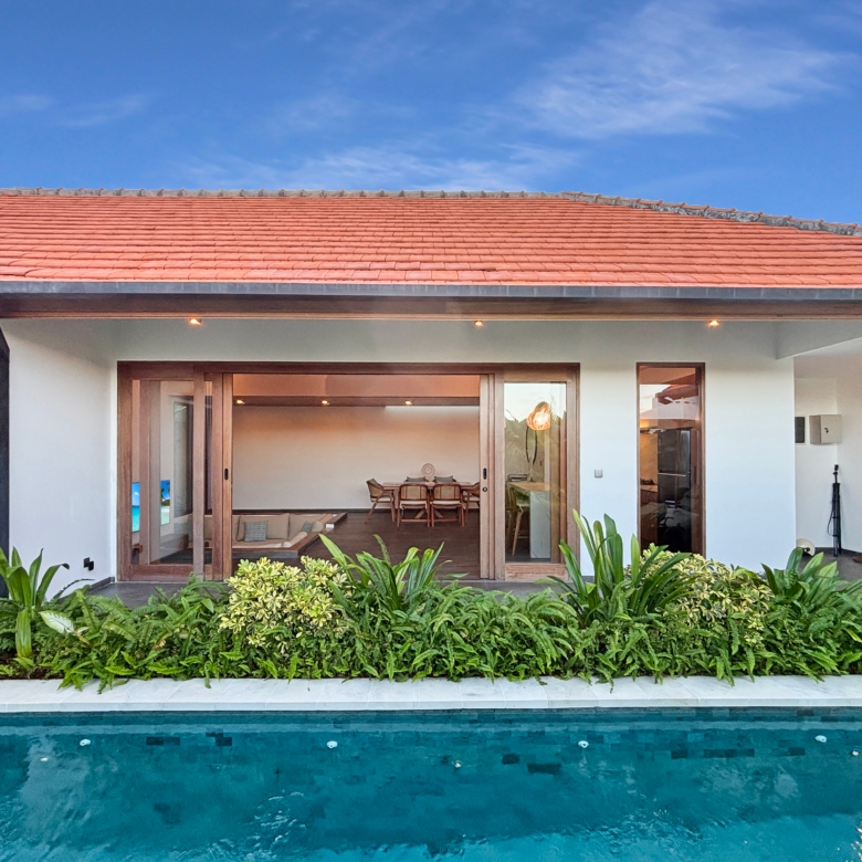 Modern villa with a red tile roof, large glass doors, and a pool in the foreground surrounded by greenery.