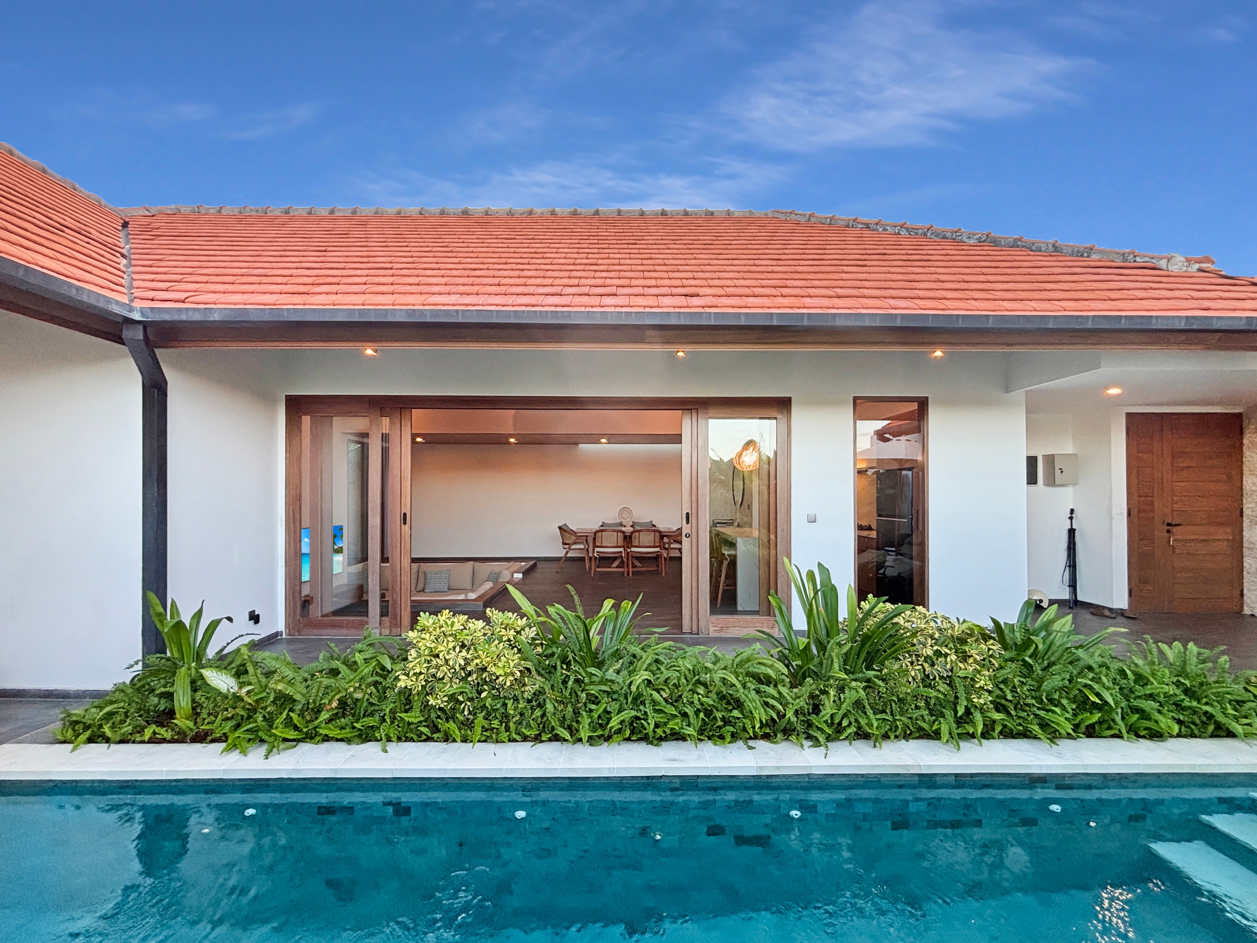 5464bc9fdf436fa7584c83cb33007fb69d6cae1f Modern villa with a red tile roof, large glass doors, and a pool in the foreground surrounded by greenery.