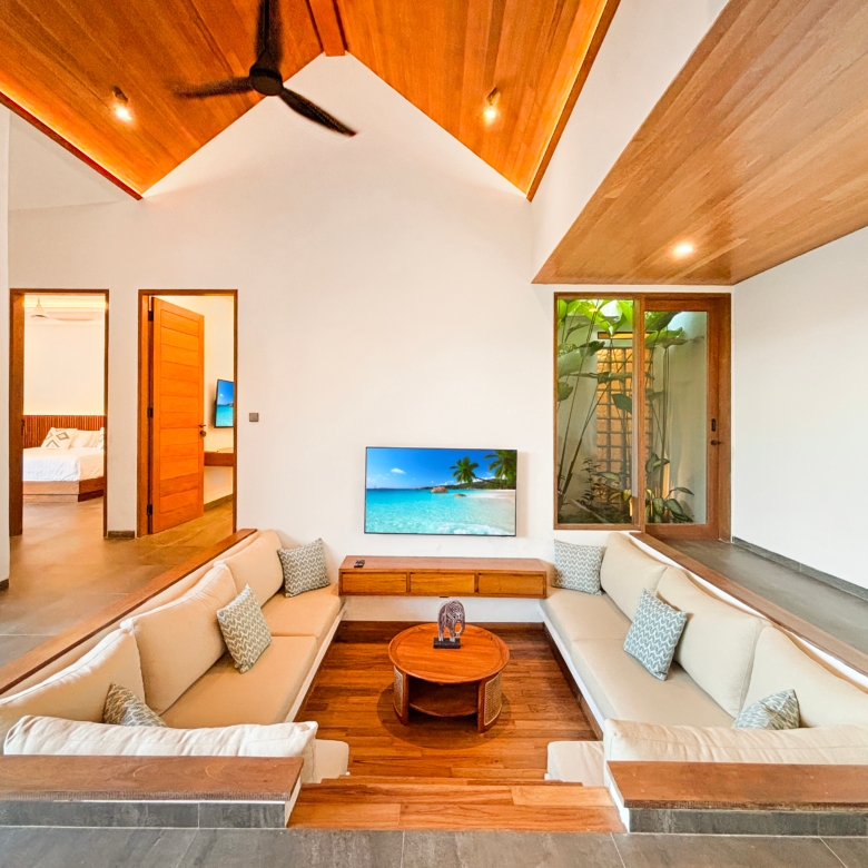 Open living room with beige sectional sofas around a wooden coffee table; wall-mounted TV shows a tropical beach scene.