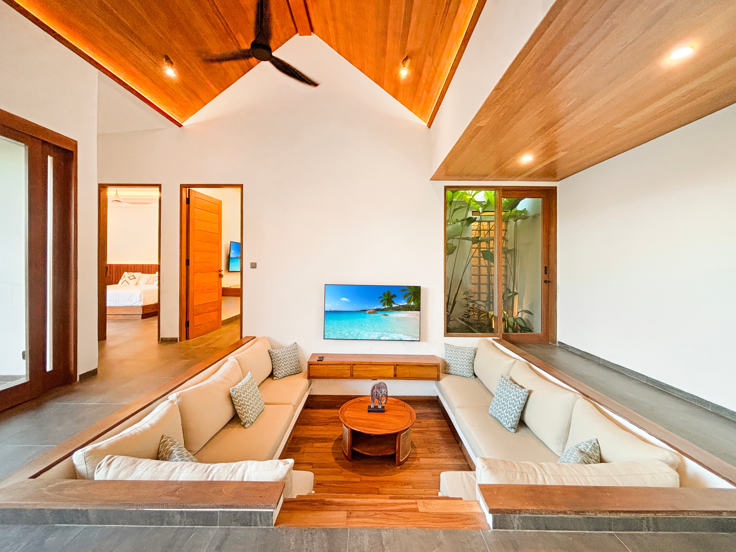 5641e77ffff51f99b4949ee6c17af8b56ed974a9 Open living room with beige sectional sofas around a wooden coffee table; wall-mounted TV shows a tropical beach scene.