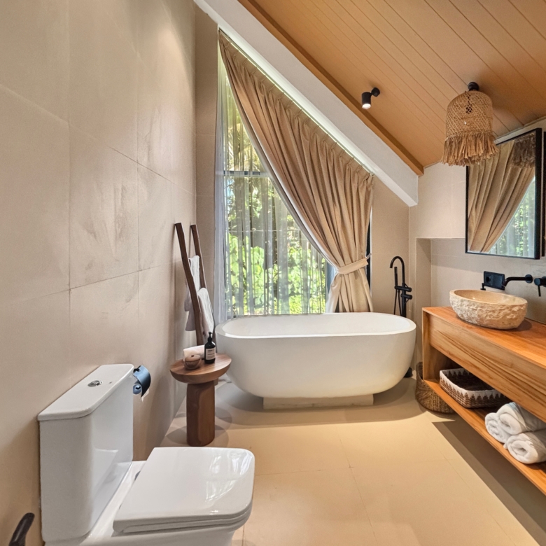 Modern bathroom with a white freestanding bathtub under a slanted wooden ceiling and beige curtains by a window outlet lighted by a wall lamp.