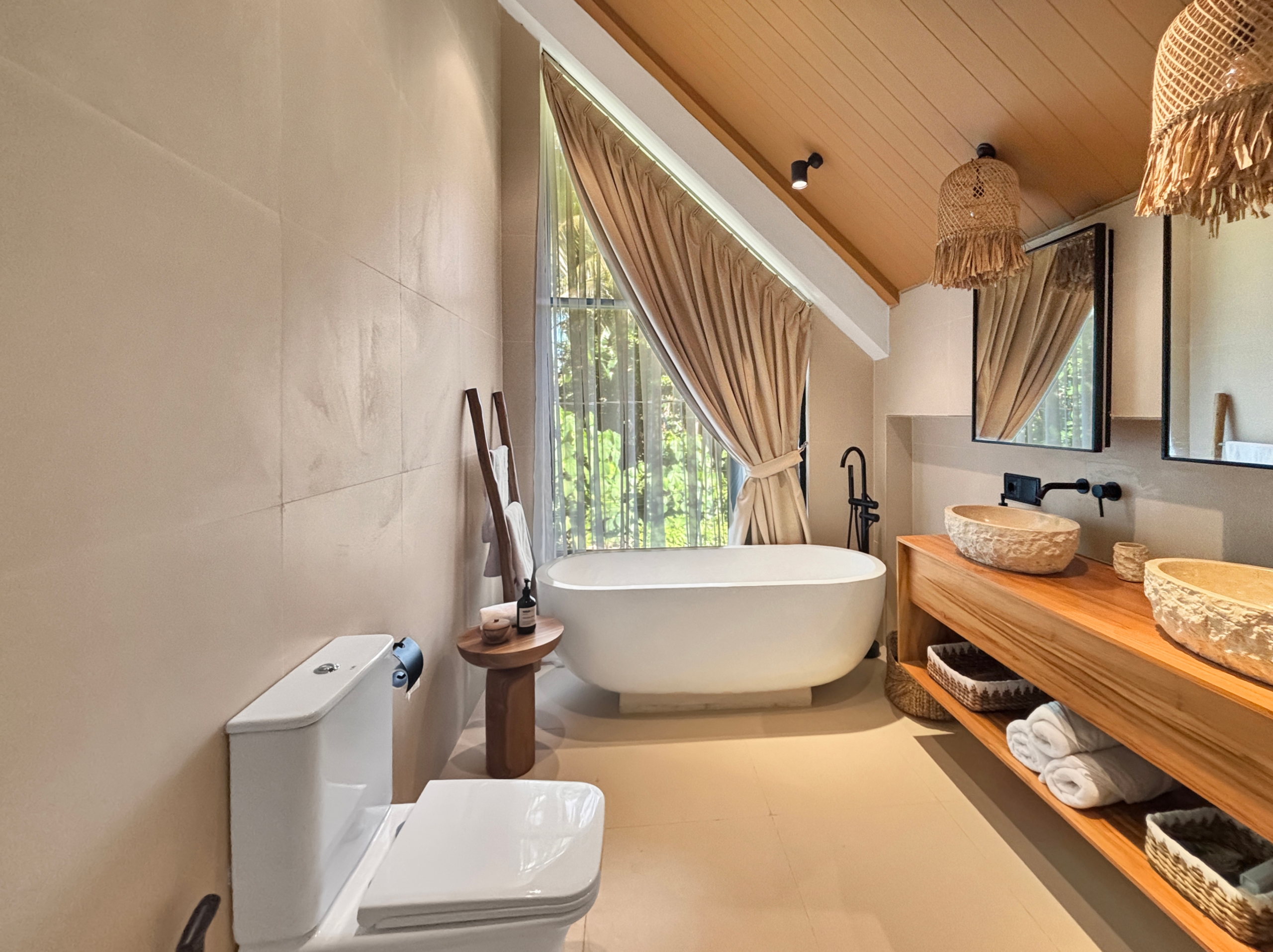 5818c7330aa678b25c6abf6a4db1129815a86ef2 Modern bathroom with a white freestanding bathtub under a slanted wooden ceiling and beige curtains by a window outlet lighted by a wall lamp.