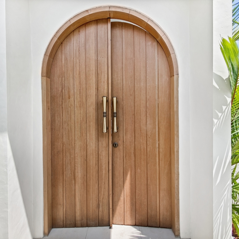 Double wooden arched doors with brass handles set in a white building, palm fronds to the right.