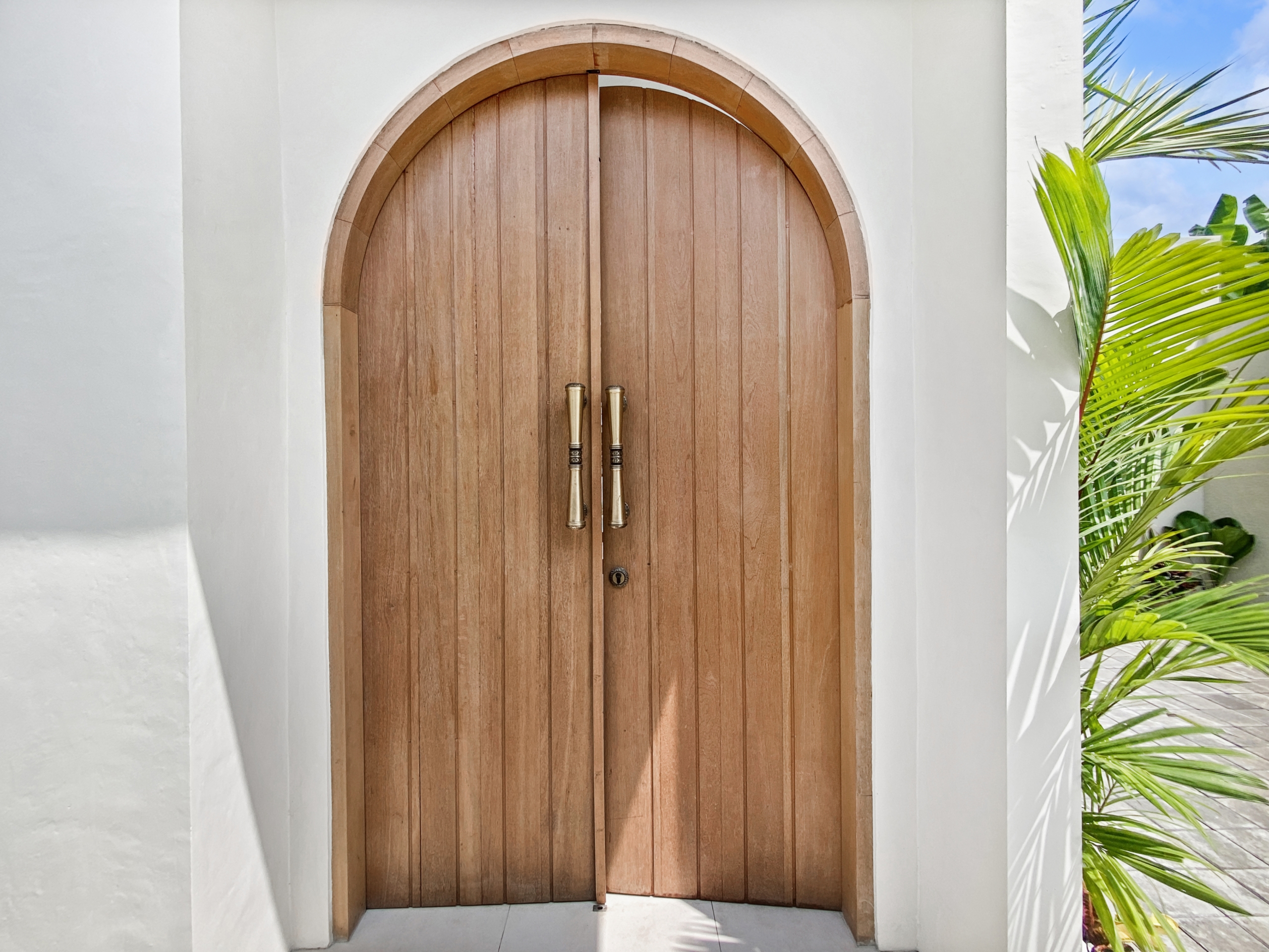 5ac1e34f00fe6ab178c8f8b63bc2d4b3a169cae3_2281-68d5cfade11e4 Double wooden arched doors with brass handles set in a white building, palm fronds to the right.