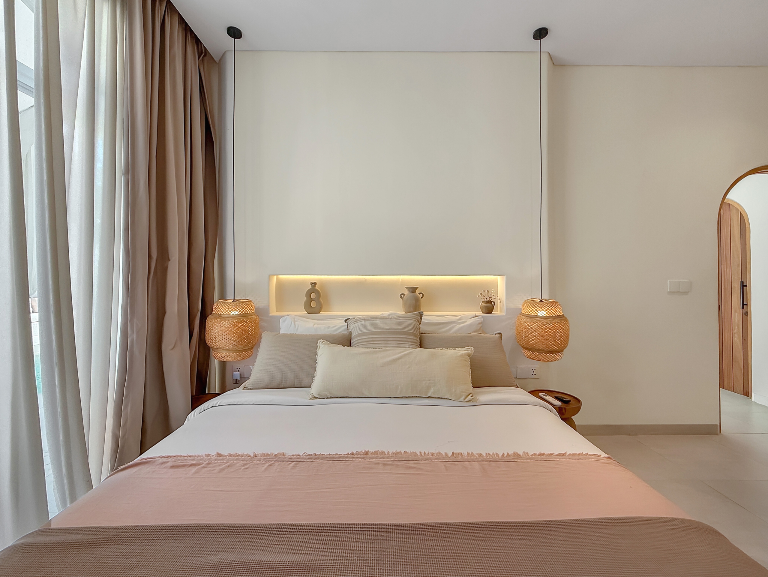 5b1a53aa635bcfcb373a9794a76ffd1570e3e02e Cozy hotel bedroom with a large bed, beige linens, woven pendant lamps, and pale curtains on the left side.