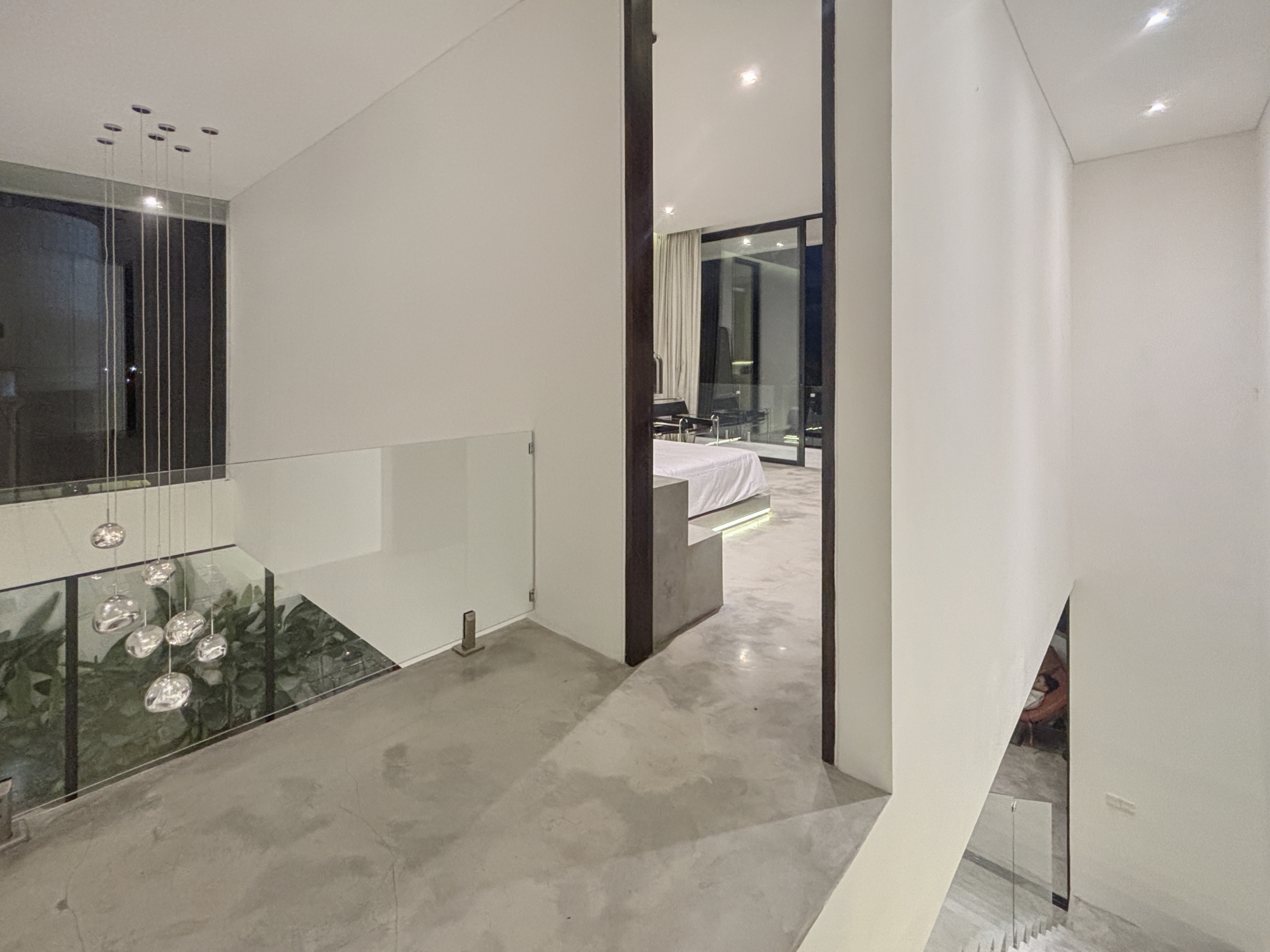 5ef118b22cd15f75484071b866eb8cf5a064d9ad_0893-6a05be0786b92 Modern interior with glass railing and hanging spherical pendant lights, leading to a doorway showing a bedroom beyond.