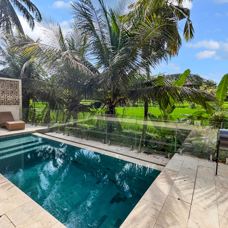 Poolside area with a rectangular turquoise pool, beige stone deck, glass railing, and surrounding tropical palm trees under a bright blue sky.