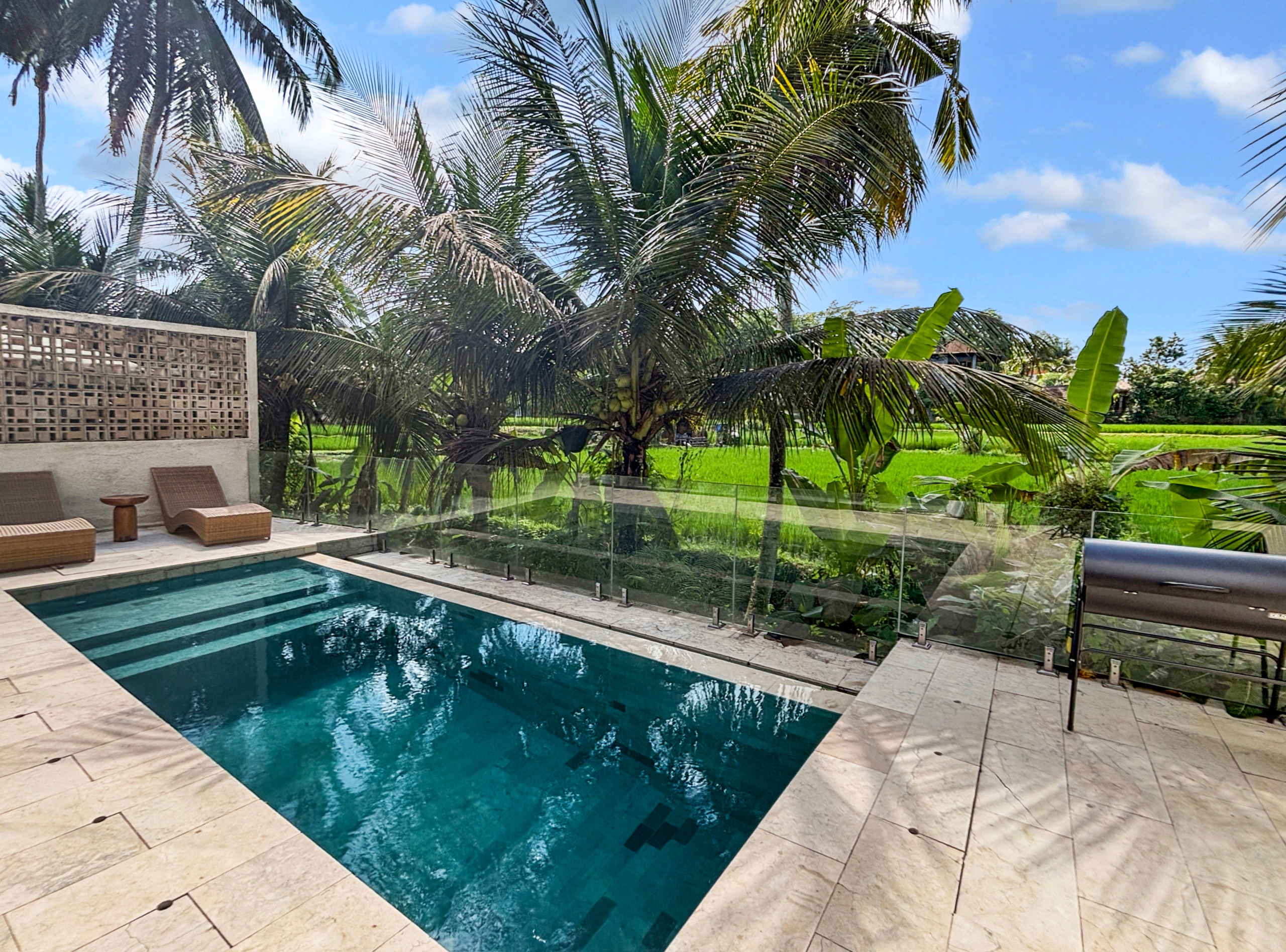 60066ba62b1adf7cca0890bf723da42ae3863800 Poolside area with a rectangular turquoise pool, beige stone deck, glass railing, and surrounding tropical palm trees under a bright blue sky.