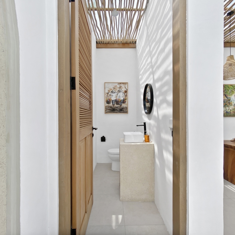 Narrow hallway opening into a bathroom with a white sink on a stone pedestal, toilet, wooden louvered door, and framed artwork on white walls.