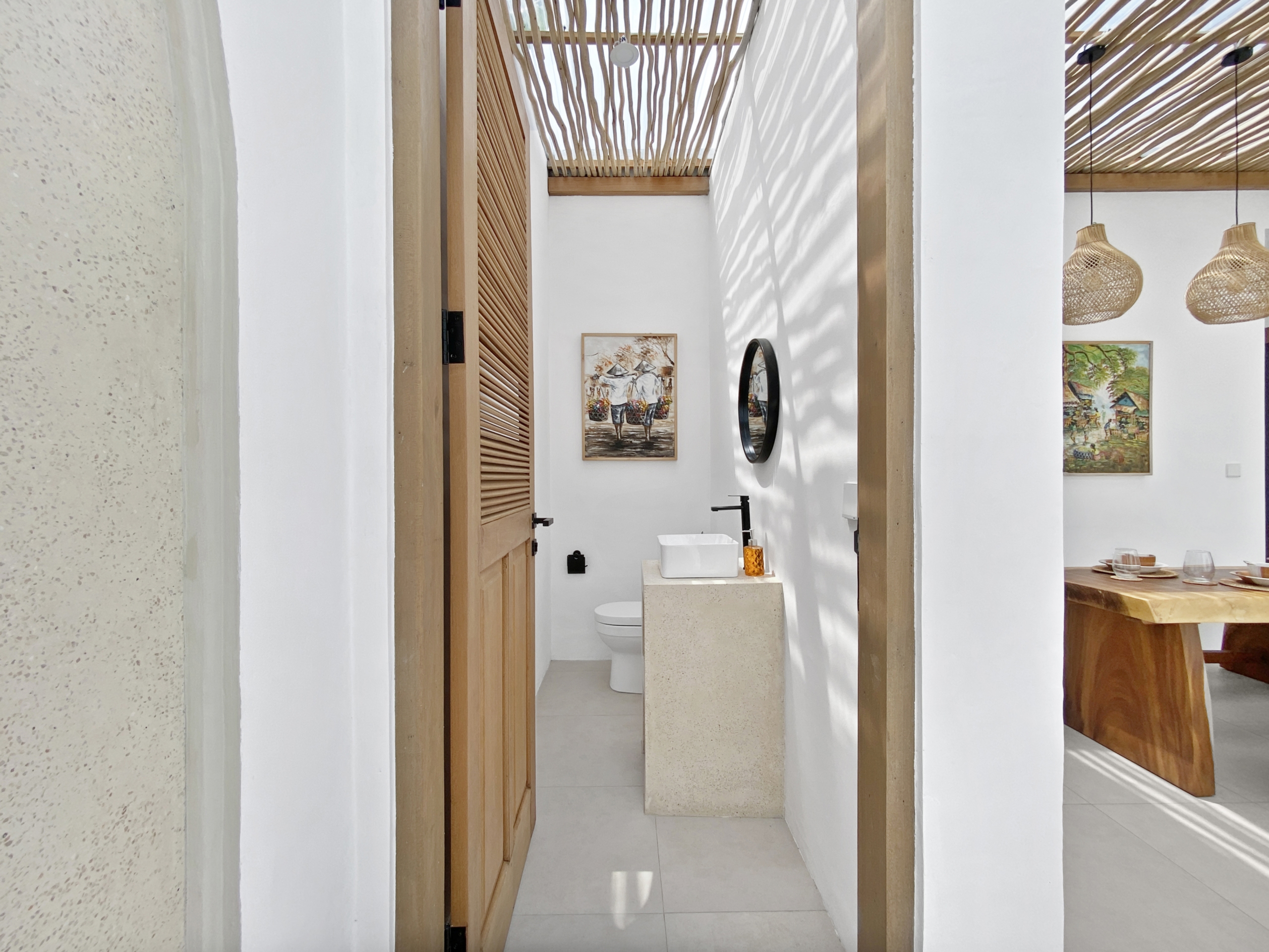 621d986d6378fccea11799c3a6e9e71c3e930741_2310-68d5cfb1a4256 Narrow hallway opening into a bathroom with a white sink on a stone pedestal, toilet, wooden louvered door, and framed artwork on white walls.