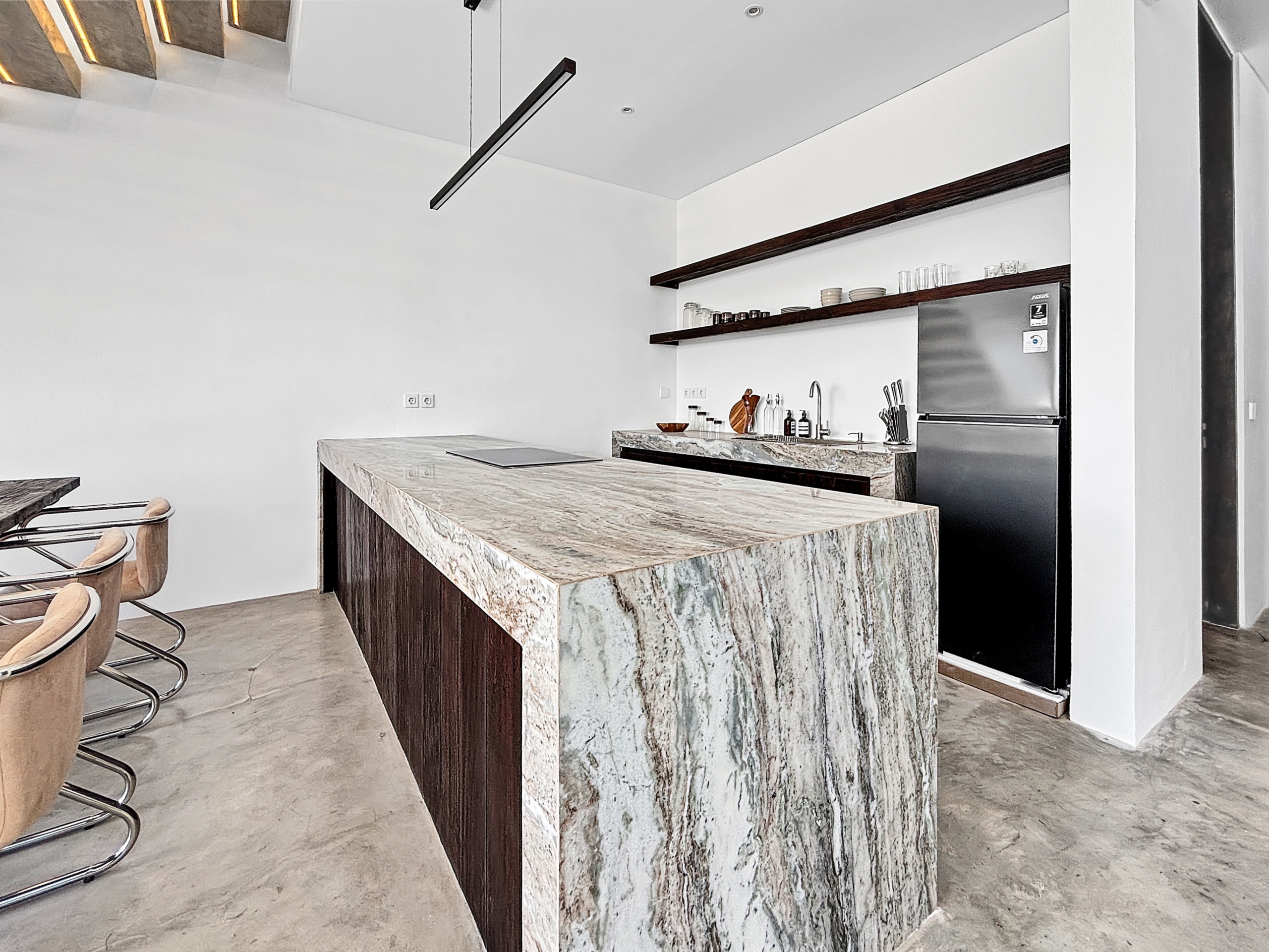 62f56ba2b8213ebade0ccd68fe5dc43e79b02b9b_6806-6a1d7baeb4c00 Modern kitchen featuring a marble island, dark wood accents, open shelves with glassware, and a stainless steel refrigerator.