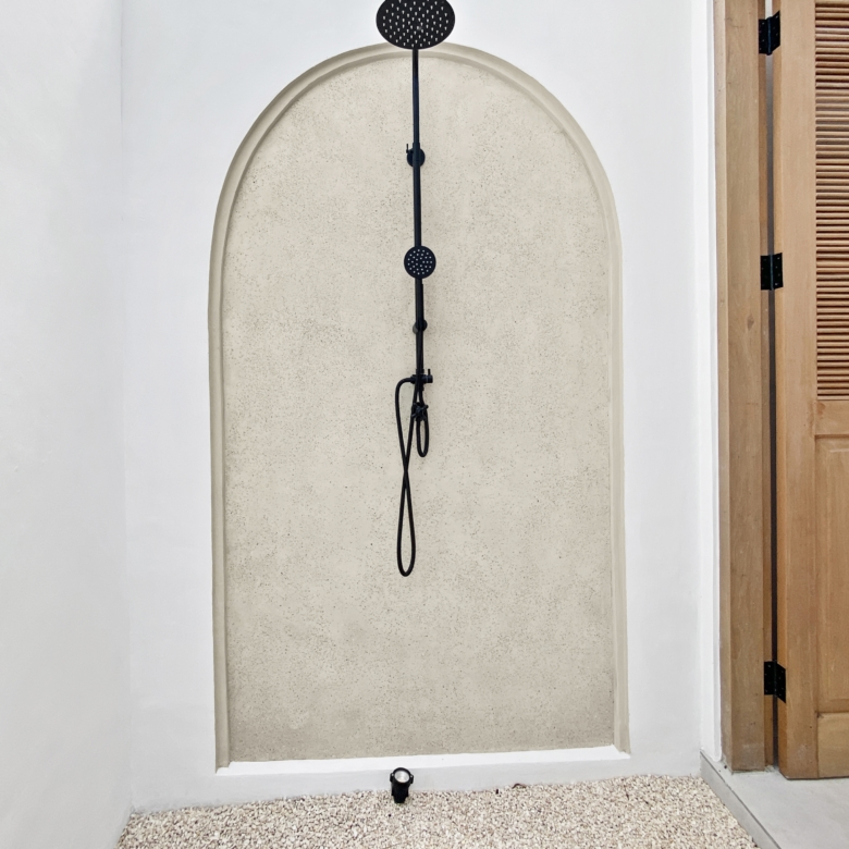 Narrow shower alcove with a vertical shower rail and two round shower heads mounted on a white wall, wood door to the right.