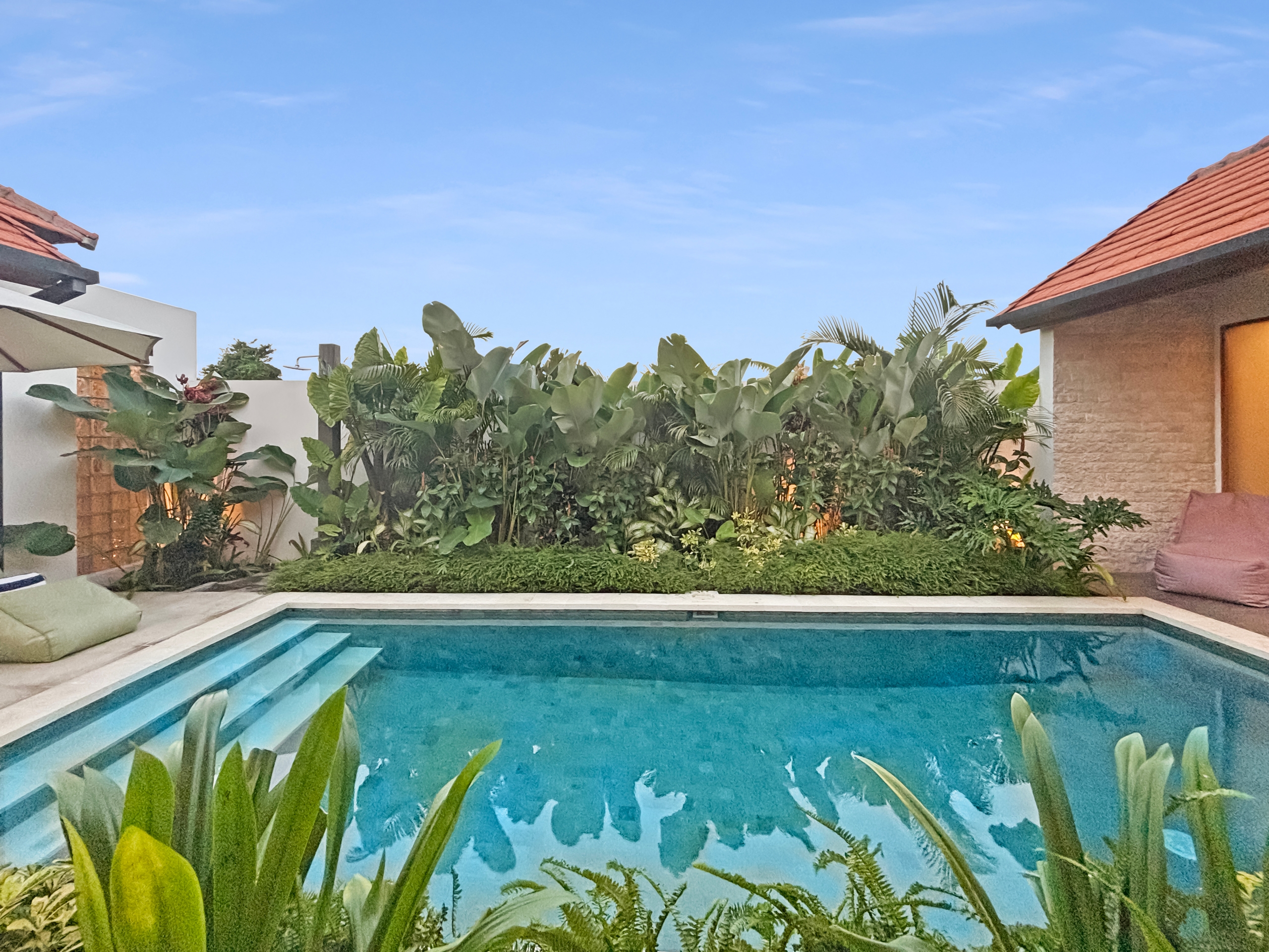 6727300ae36d4990fc913d44826ddc9d1523b249 Backyard pool area with a rectangular swimming pool, white walls, and lush tropical plants, a red-tiled roof on the right building visible.