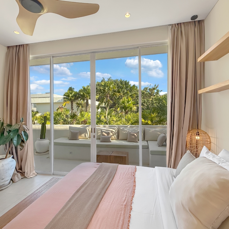 Bright bedroom with sliding glass doors opening to a sunny balcony, beige curtains, and several potted plants inside.